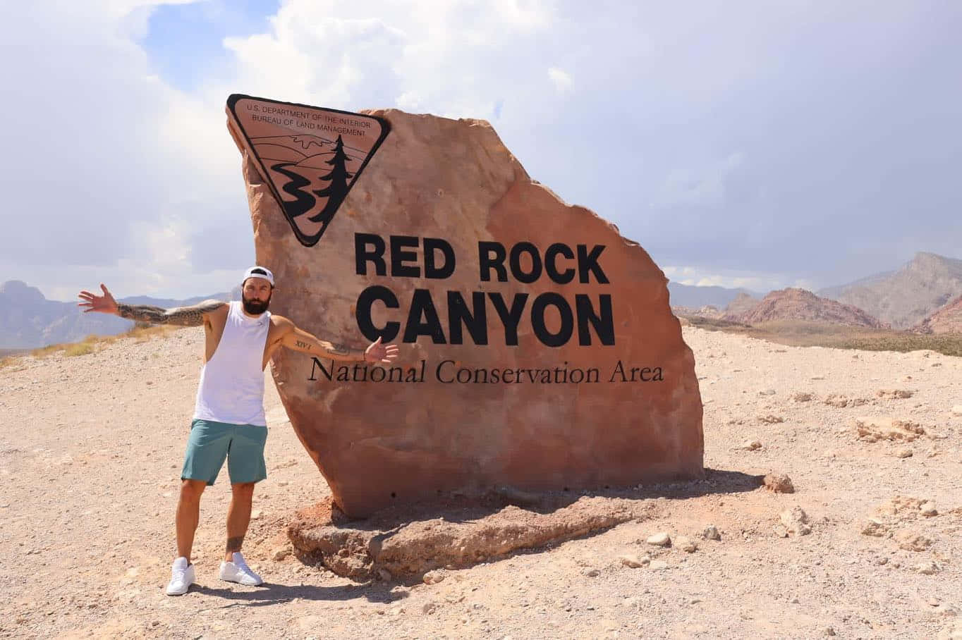 Red Rock 桌布 壁紙 — 一名男子張開雙臂，自豪地站在紅岩峽谷國家保護區的大型紅色岩石標誌前，天空部分多雲。