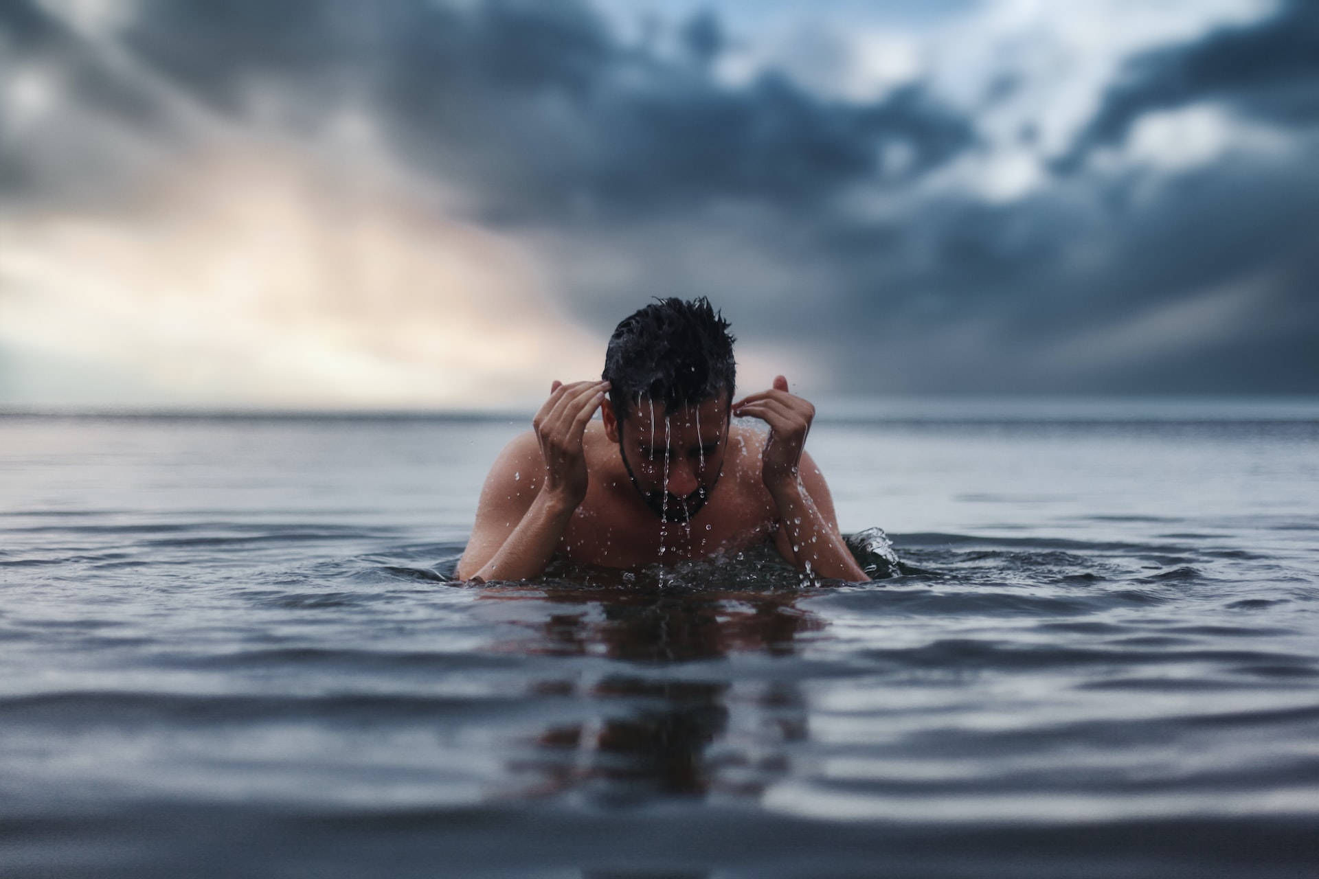 Swimming 桌布 壁紙 — 一名男子從波濤洶湧的海水中浮現，雨水從他濕透的頭髮滴落，背景是戲劇性的多雲天空。
