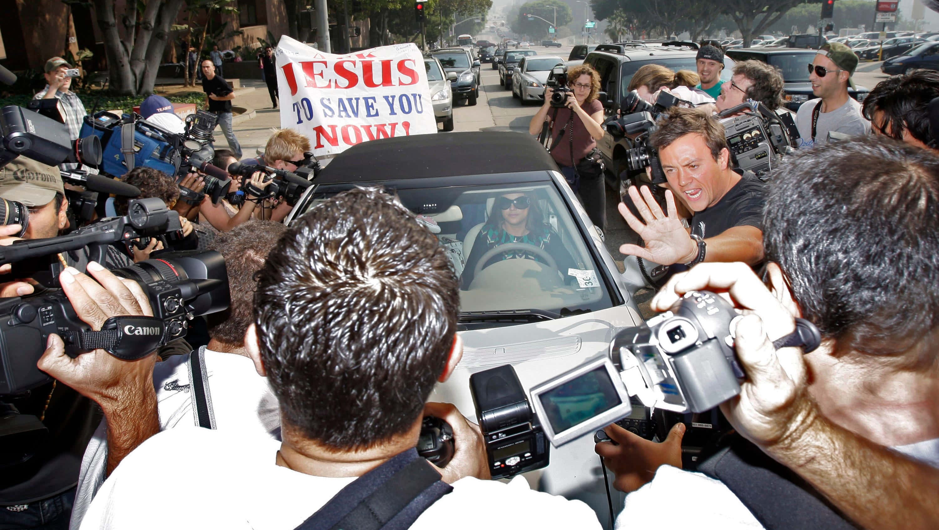 Paparazzi 桌布 壁紙 — 一名男子從車內揮手，周圍是攝影師和一名舉著「Jesus to save you now」標語的抗議者。