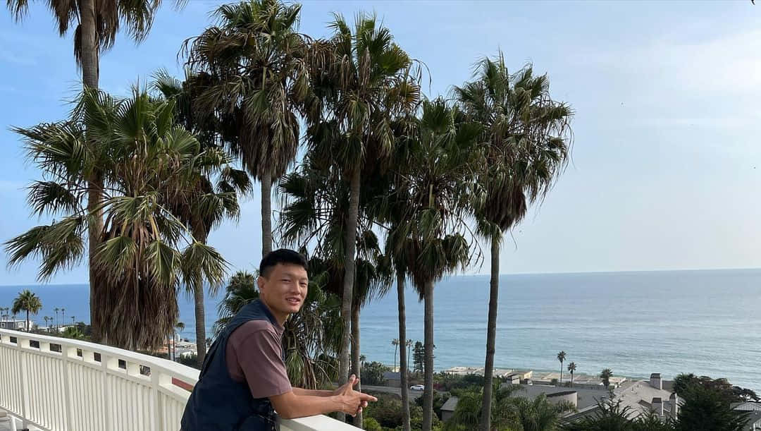 Beach Vacation 桌布 壁紙 — 一名男子微笑著靠在白色欄杆上，俯瞰著平靜的海洋，四周是高聳的棕櫚樹，頭頂是清澈的藍天。