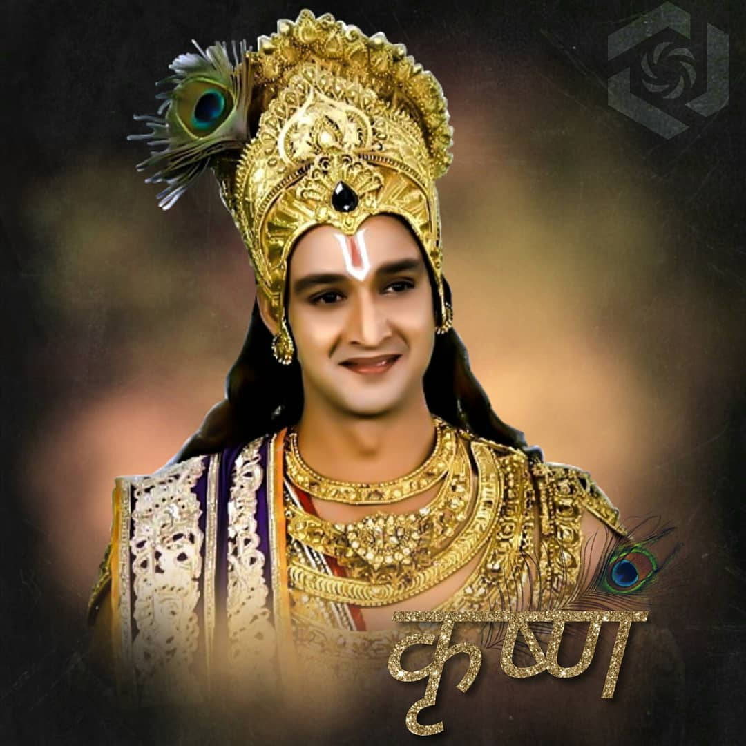 Mahabharat Krishna 桌布 壁紙 — 一名男子打扮成克里希納神，身穿華麗金色服飾，飾有孔雀羽毛和神聖蒂拉克，在黑暗模糊的背景前溫暖微笑。