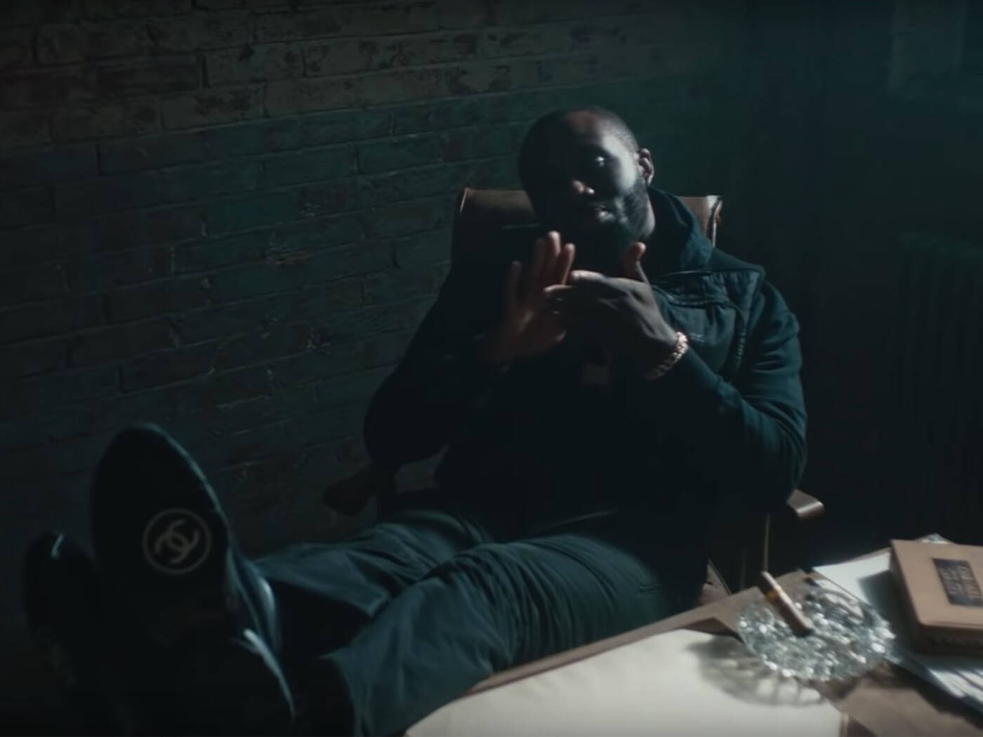 Skepta 桌布 壁紙 — 一名男子放鬆地坐在光線昏暗的磚牆房間裡，雙手比劃著好像在說話，身穿深色衣服和 Chanel 鞋子。
