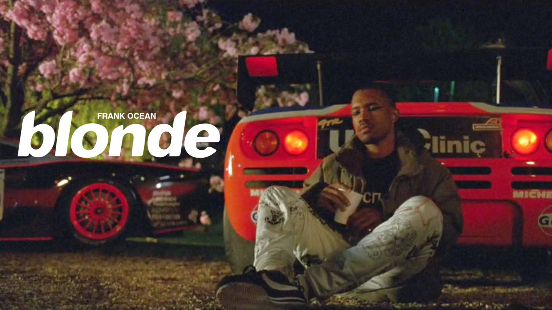 Frank Ocean 桌布 壁紙 — 一名男子晚上坐在粉紅色櫻花下的紅色賽車旁邊的地上，尾燈閃閃發光。