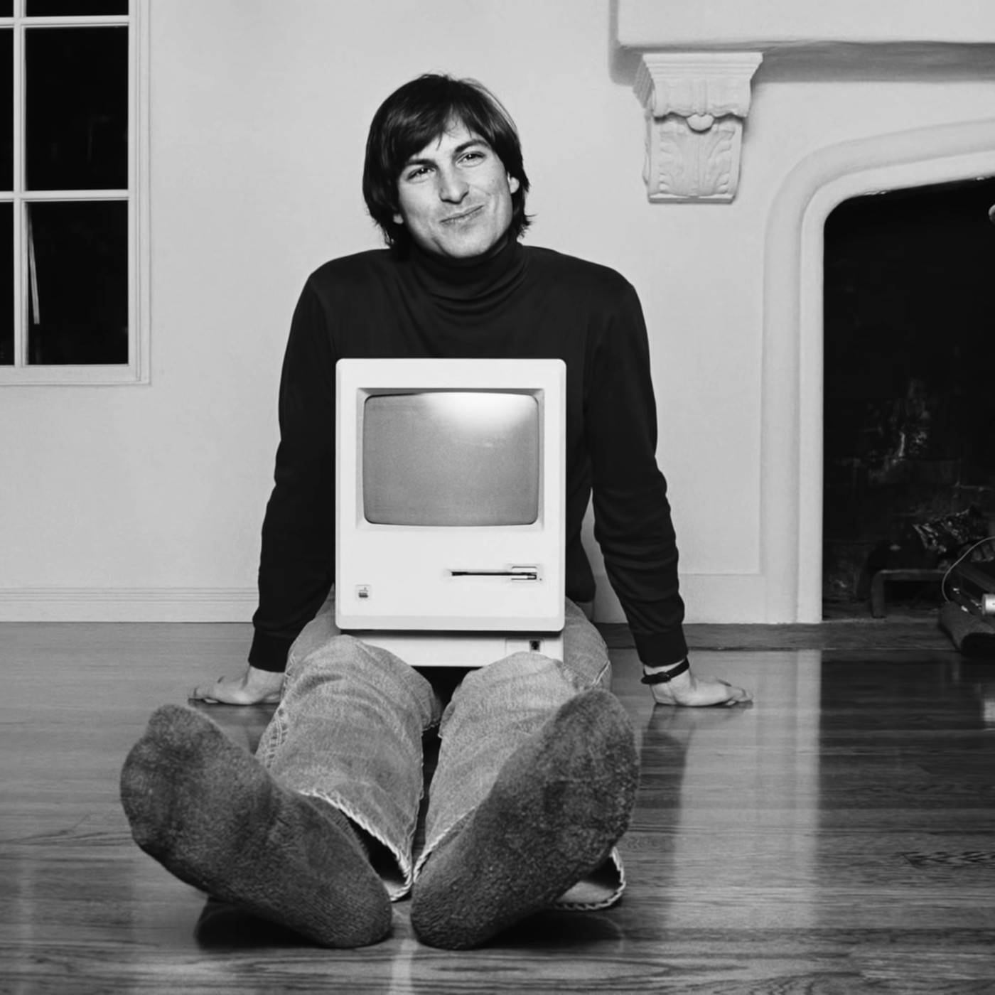 Rich 桌布 壁紙 — 一名男子盤腿坐在硬木地板上，懷裡放著一部復古Apple電腦，在單色室內環境中對著鏡頭微笑。