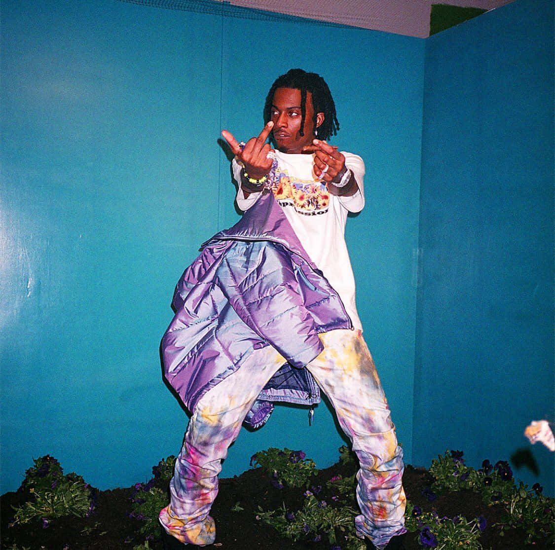 playboi carti pfp 桌布 壁紙 — 一名男子穿著鮮豔的紮染褲和紫色羽絨外套，在純藍色牆壁前擺姿勢，腳邊有紫色花朵。