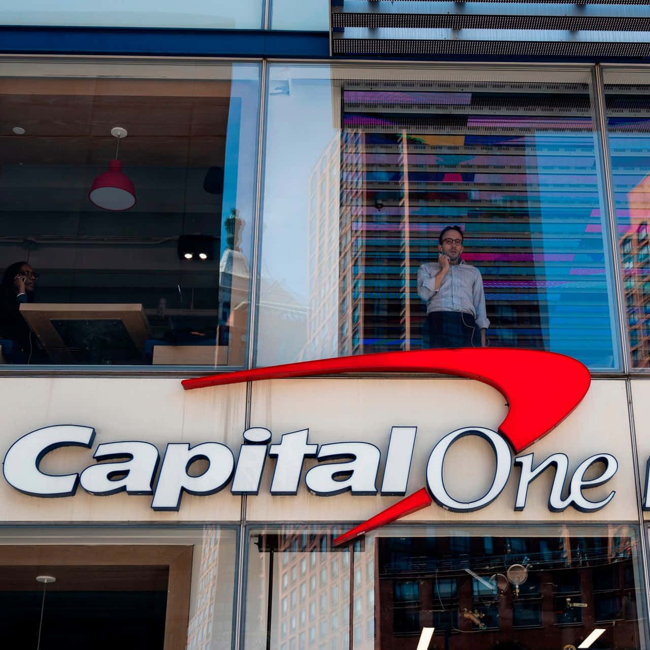 Man 桌布 壁紙 — 一名男子站在Capital One辦公室的窗戶前，窗戶反射出城市摩天大樓和繽紛燈光，背景是明亮的白天。