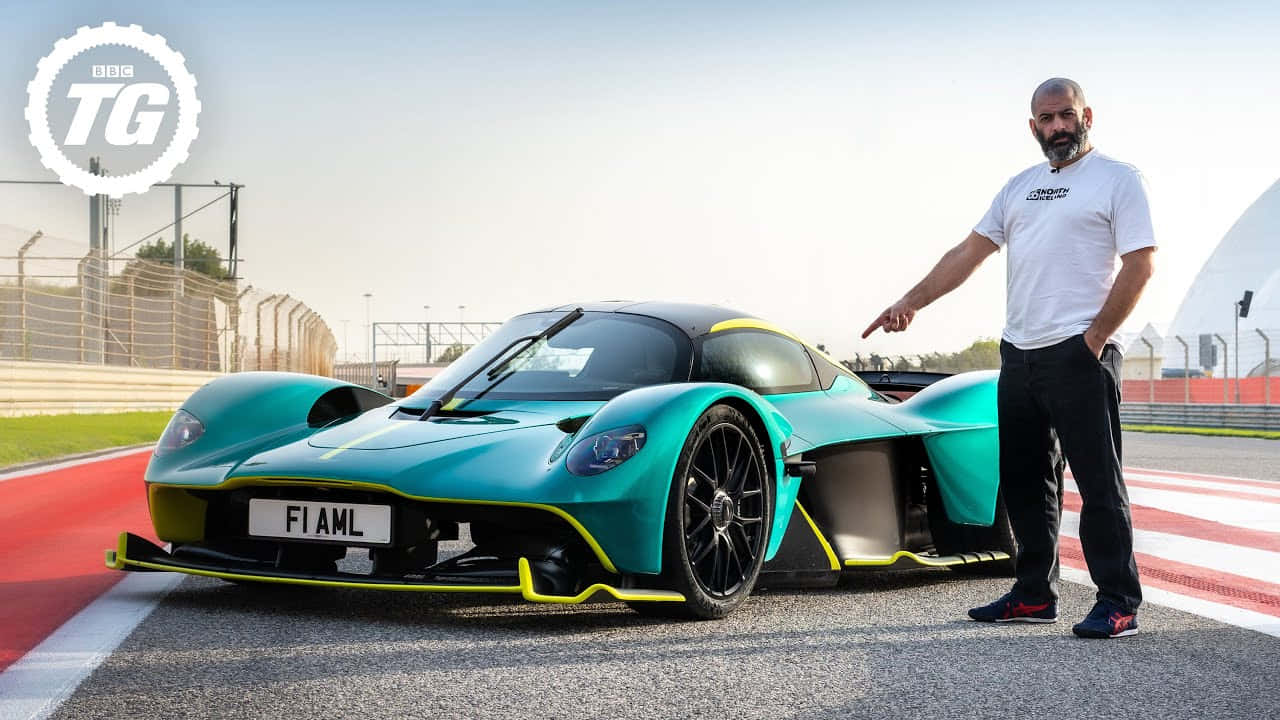 奧斯頓馬丁 桌布 壁紙 — 一名男子站在一台青綠色和黃色的Aston Martin Valkyrie旁邊，賽道上有紅色和白色的路緣石，天空晴朗。