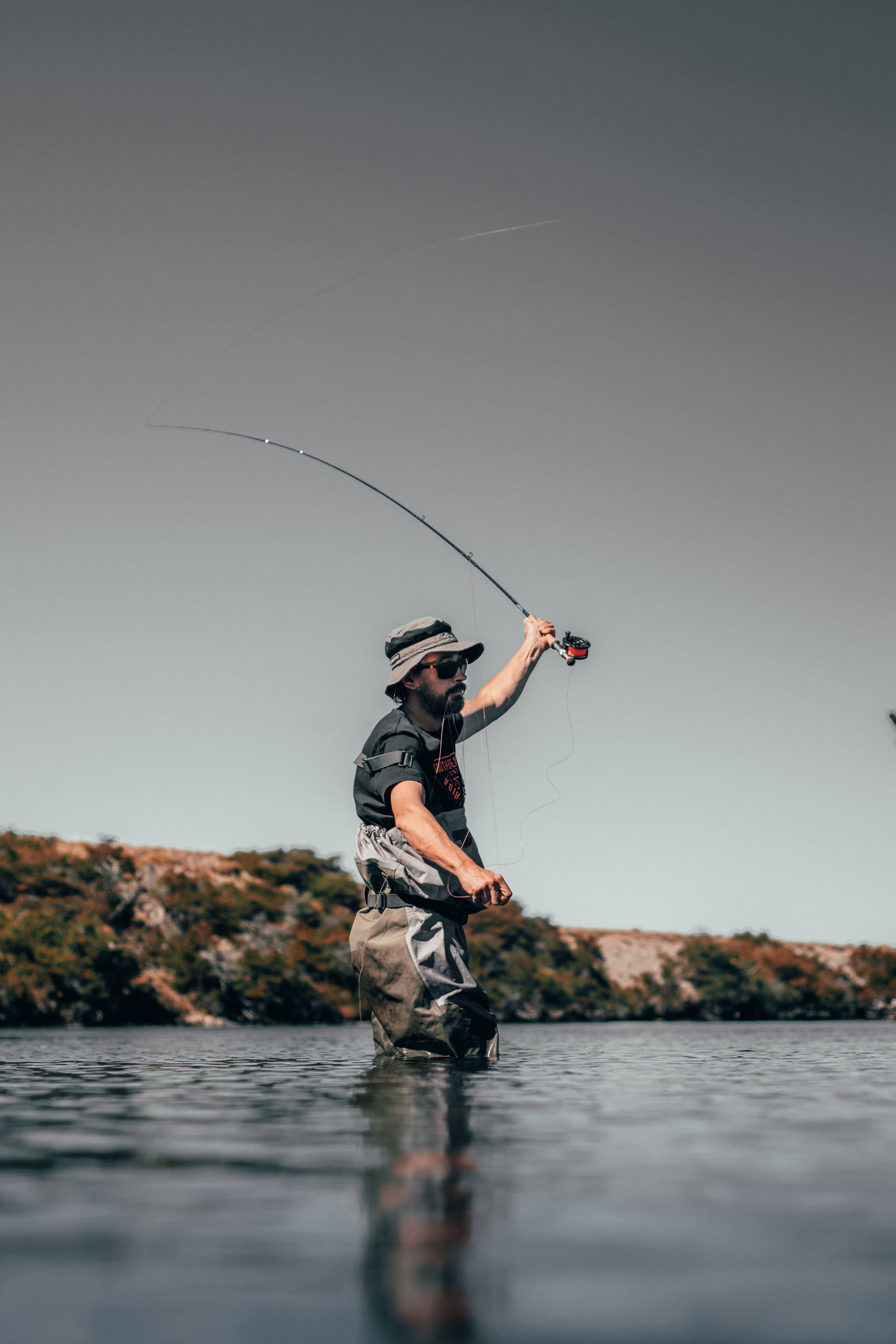 Bass Fishing 桌布 壁紙 — 一名男子站在及膝的平靜水中，向著暗淡的天空拋出釣竿，遠處有岩石海岸。