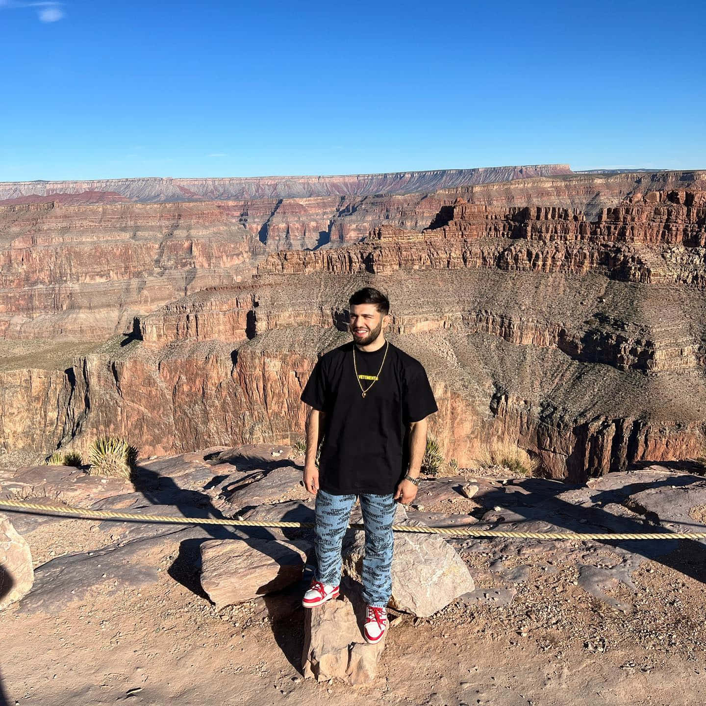  壁紙 — 一名男子站在大峽谷的岩石觀景點上微笑，天空清澈湛藍，深紅色的岩層延伸至遠方。
