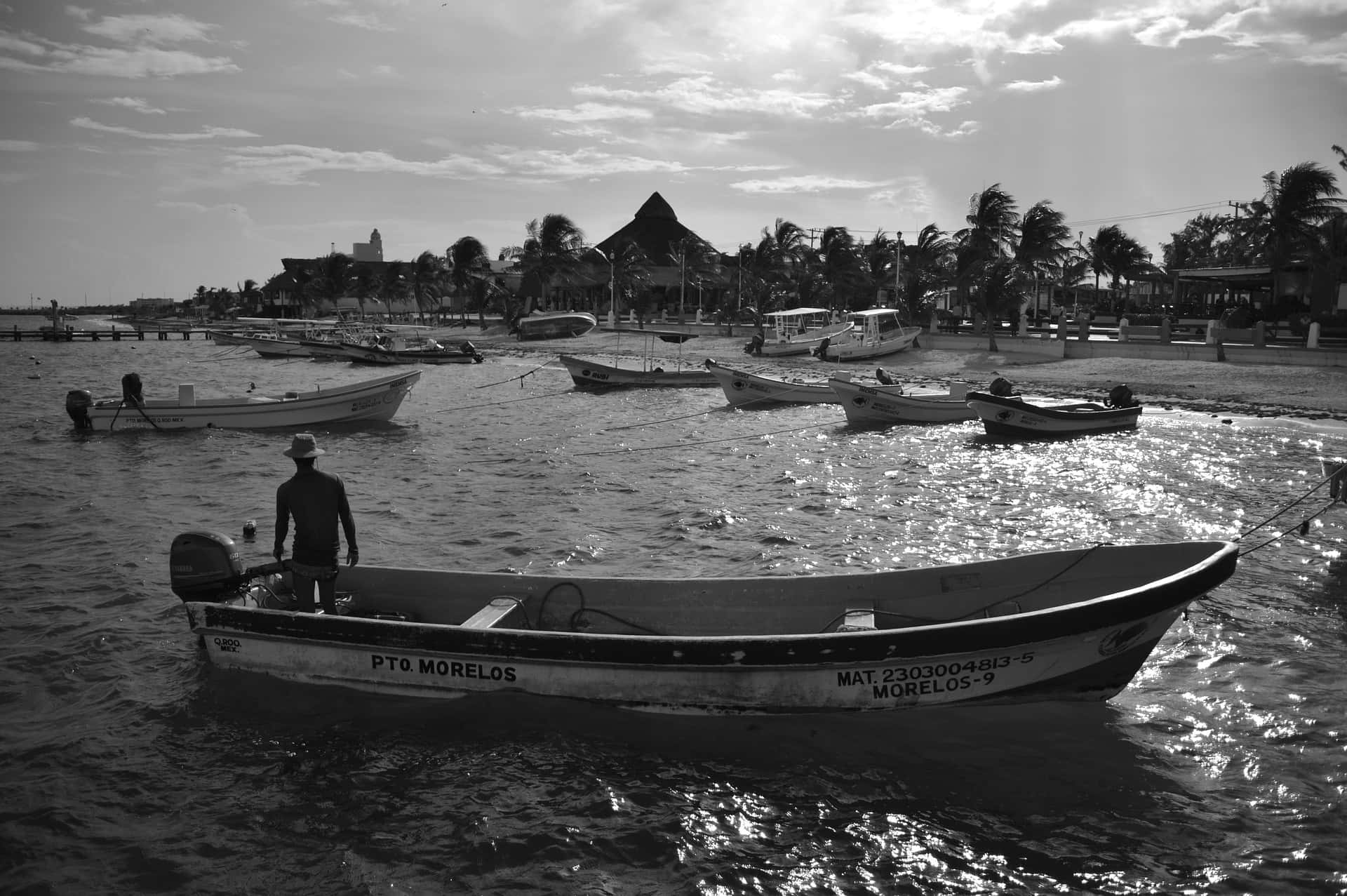 Cancun 桌布 壁紙 — 一名男子站在小型機動船上，水面平靜，背景有其他船隻和棕櫚樹，天空多雲。