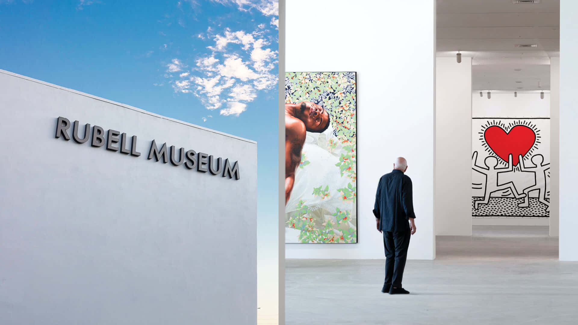 Museum 桌布 壁紙 — 一名男子站在明亮的白色畫廊中，欣賞充滿活力的藝術品，包括一幅色彩繽紛的肖像畫和基思·哈林的心形作品，背景是藍天。