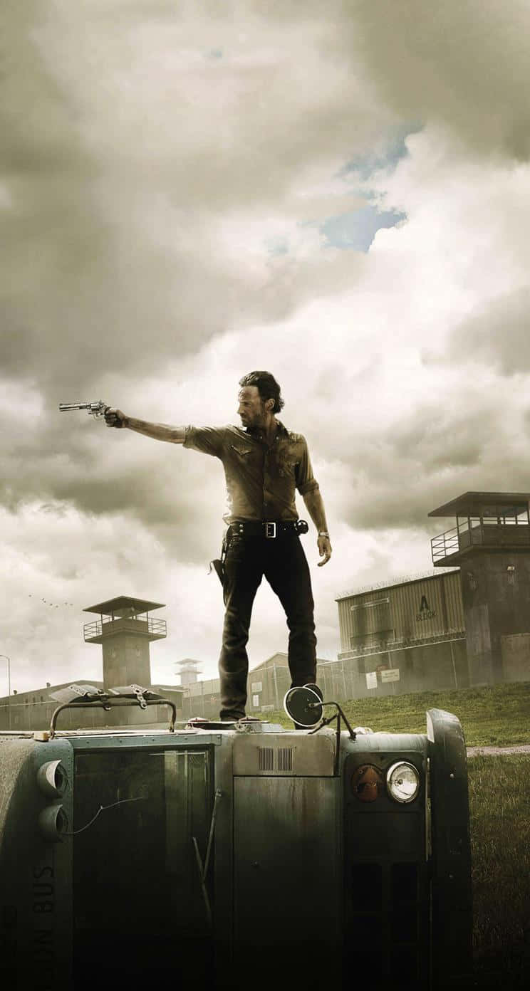 The Walking Dead iPhone 桌布 壁紙 — 一名男子站在翻覆的巴士頂上，用手槍瞄準多雲的天空，背景有監獄塔樓。