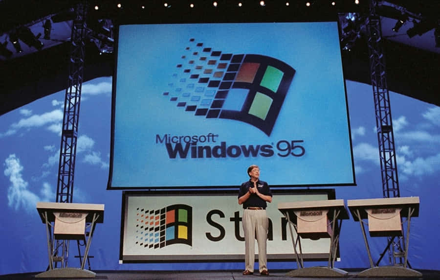 Windows 95 動態圖 桌布 壁紙 — 一名男子站在舞台上，巨型螢幕顯示著經典的Microsoft Windows 95標誌，背景是藍天。