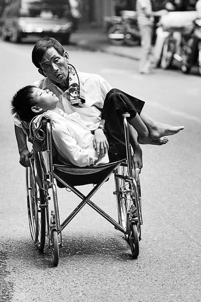 emotion 桌布 壁紙 — 一名男子背著孩子，同時推著輪椅沿街前行，黑白照片。