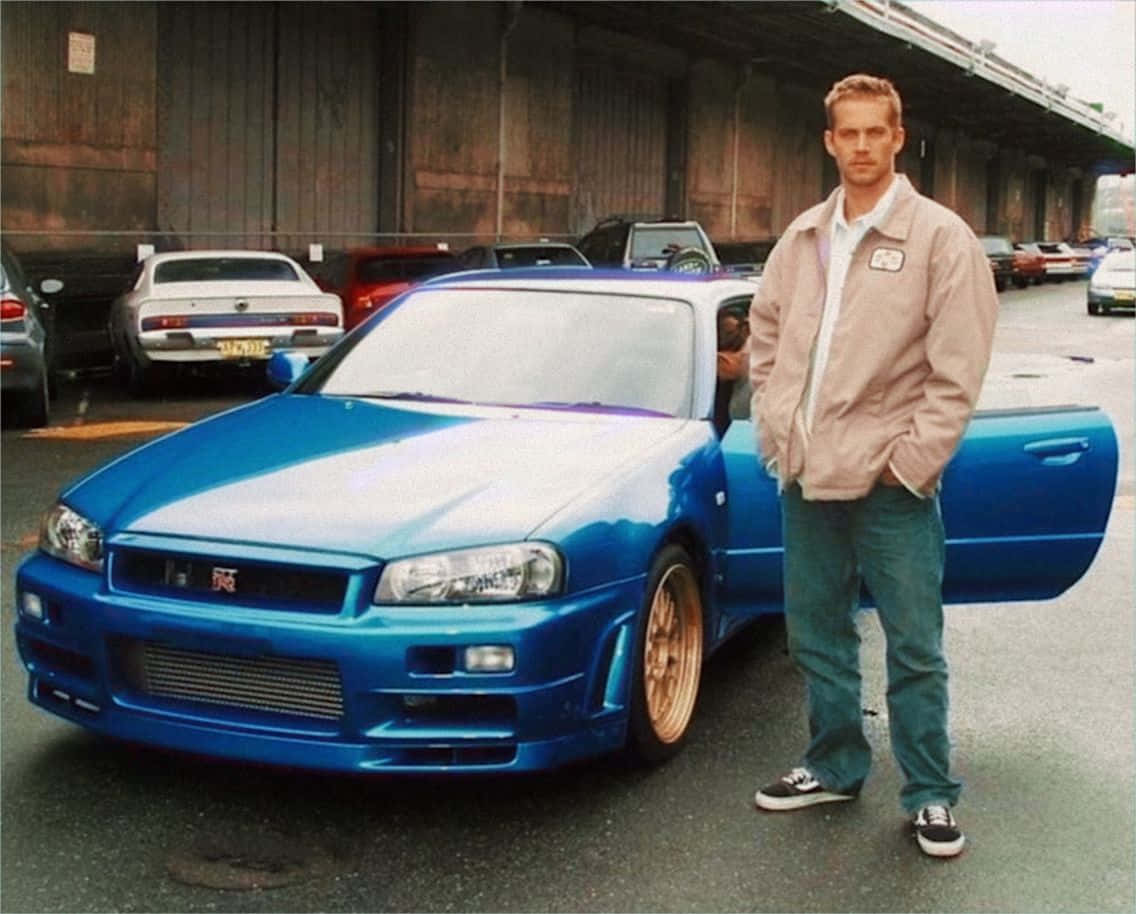 Paul Walker Skyline 桌布 壁紙 — 一名男子自信地站在一輛鮮藍色的日產Skyline R34旁邊，天橋下有其他車輛停泊在附近。