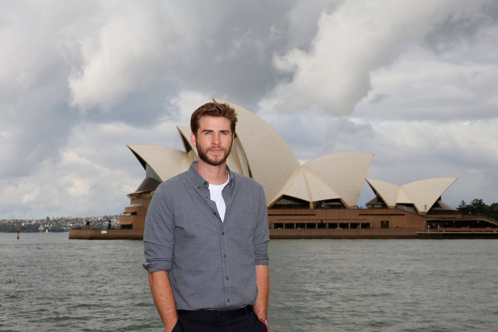 liam 桌布 壁紙 — 一名男子自信地站在標誌性的雪梨歌劇院前，天空烏雲密佈，氣氛戲劇化。