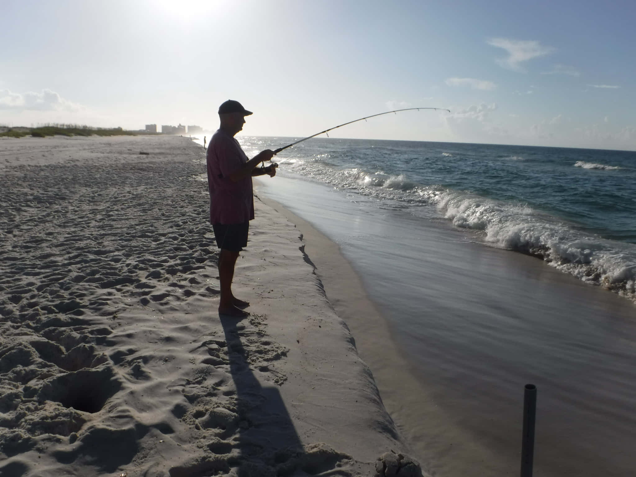 Fishing Rod 桌布 壁紙 — 一名男子赤腳站在陽光明媚的沙灘上，將釣竿拋入海浪輕拍的海中。