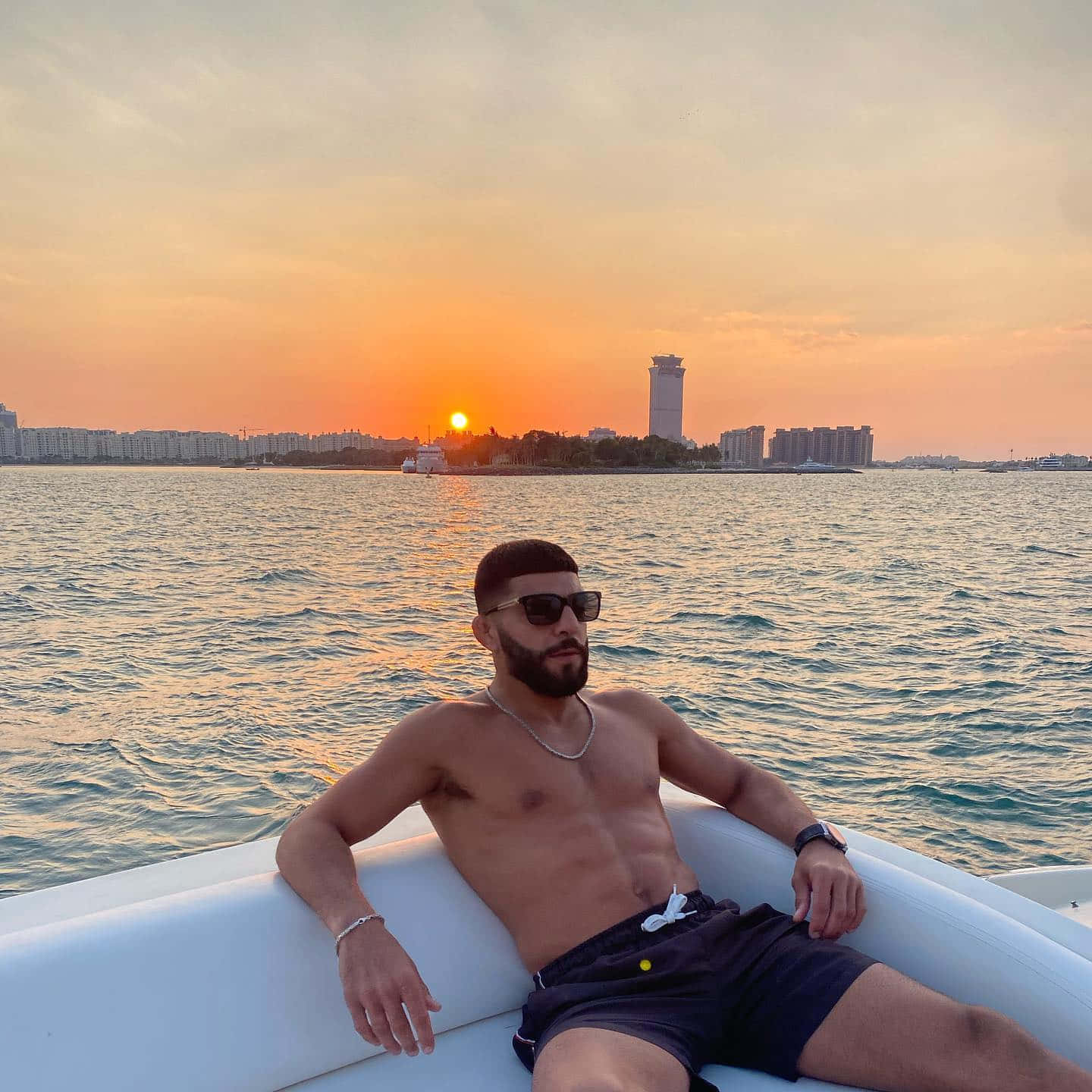 Vacation 桌布 壁紙 — 一名男子赤裸上身躺在一艘白色充氣船上，夕陽映照在平靜水面上，城市天際線倒映其中。