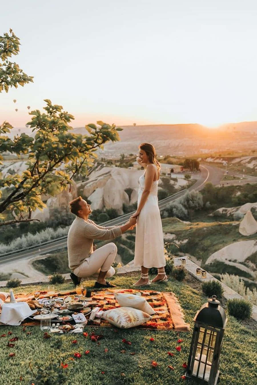 Proposal 桌布 壁紙 — 一名男子跪在圖案毯子上，向一位身穿白色連衣裙的女子求婚，地點是日落時分的草地山頂，熱氣球在獨特岩石結構上方飄浮。
