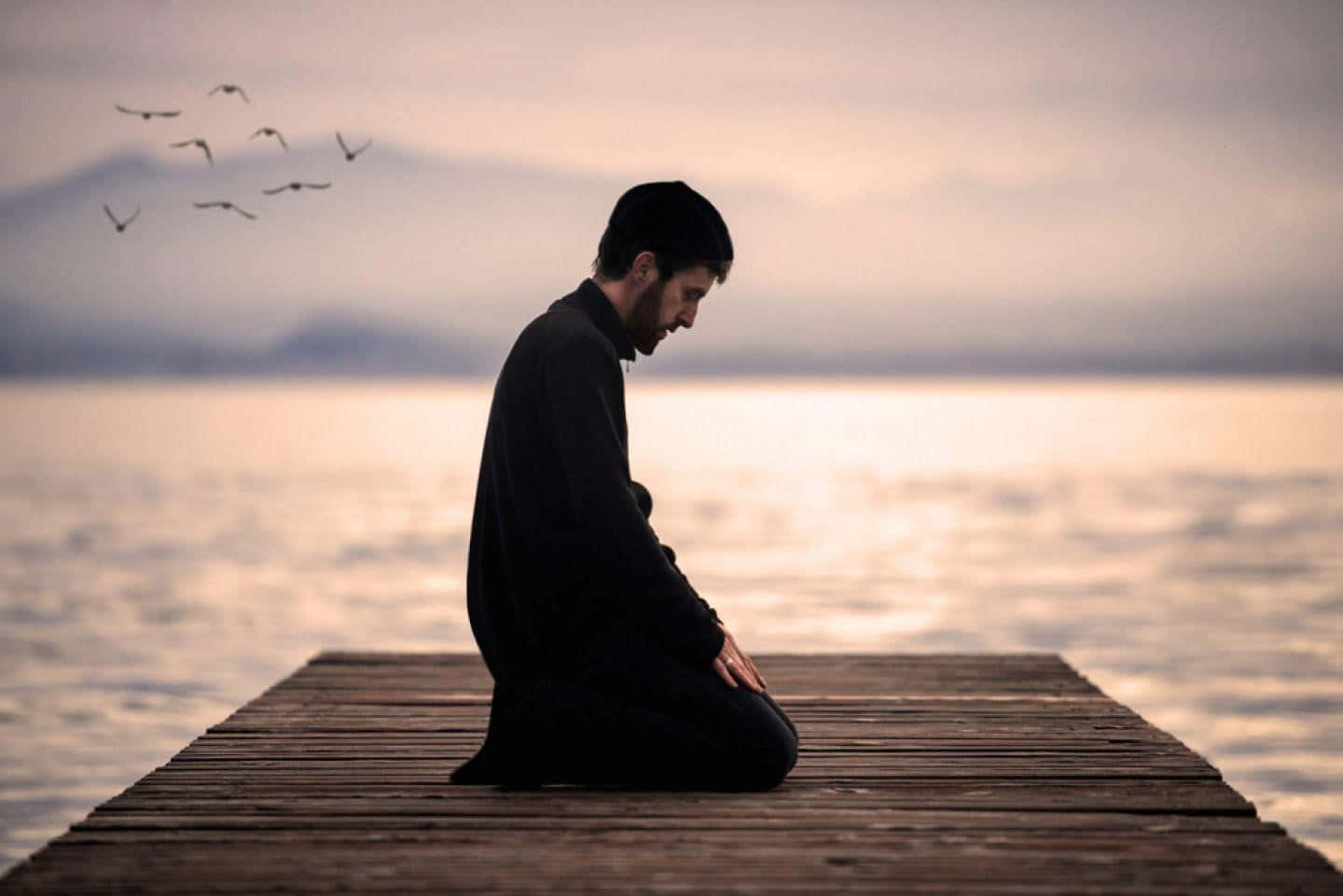 Praying 桌布 壁紙 — 一名男子跪在木碼頭上靜默沉思，鳥兒在柔和的暮色天空中飛翔。