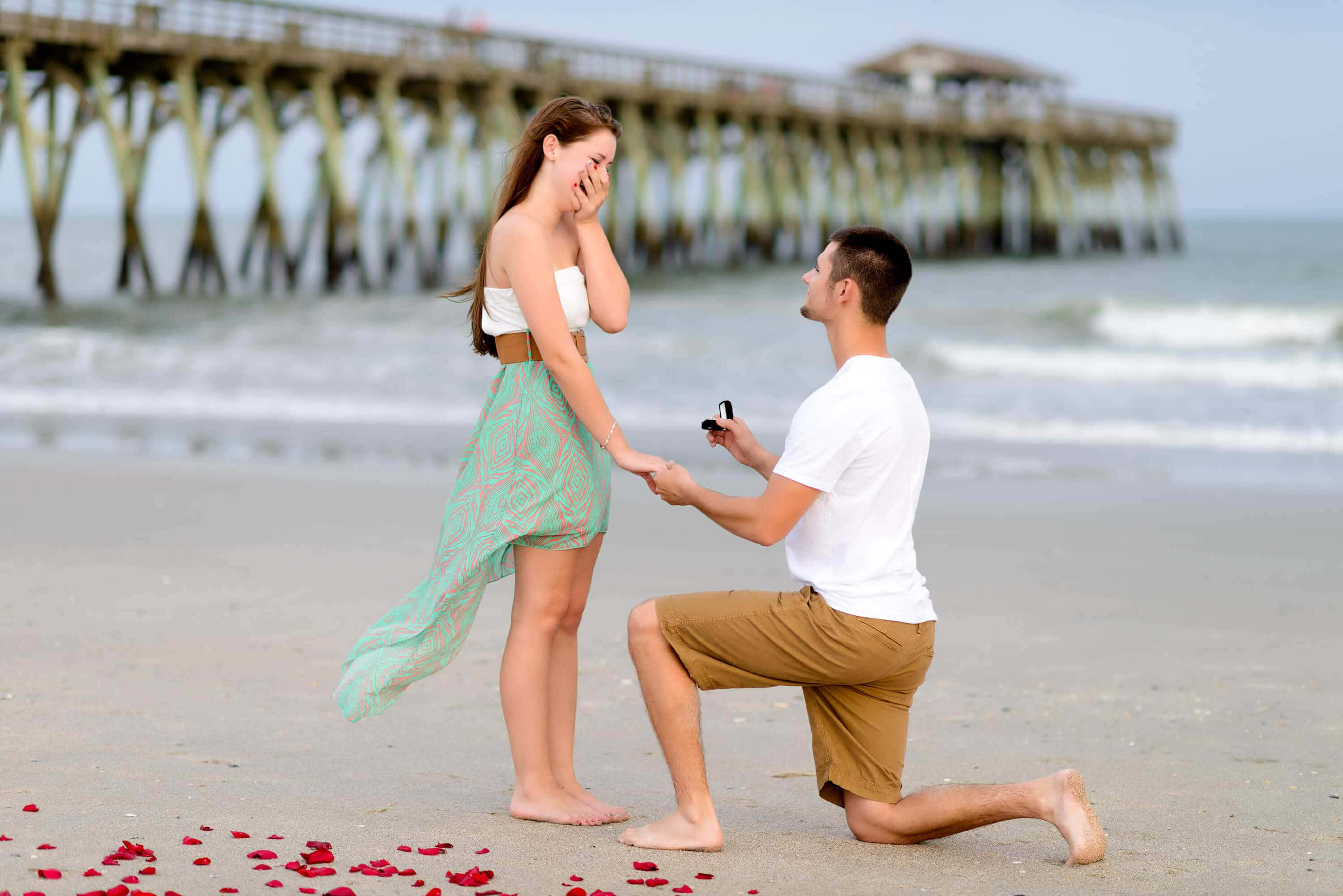 Proposal 桌布 壁紙 — 一名男子跪在沙灘上，手持戒指，女子在木製碼頭旁欣喜若狂。