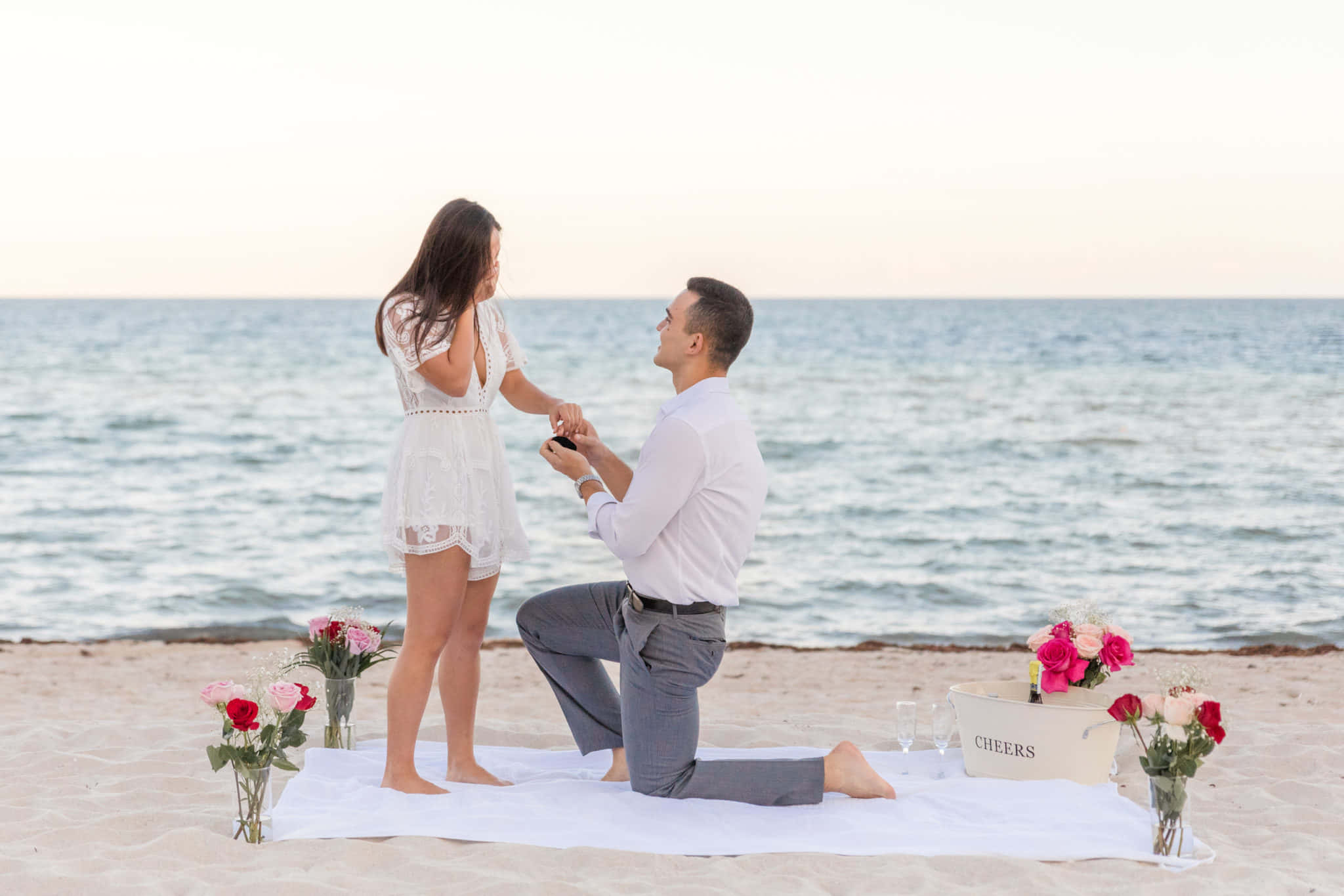 Proposal 桌布 壁紙 — 一名男子跪在沙灘毯上向一名女子求婚，夕陽下海浪在他們身後翻滾。
