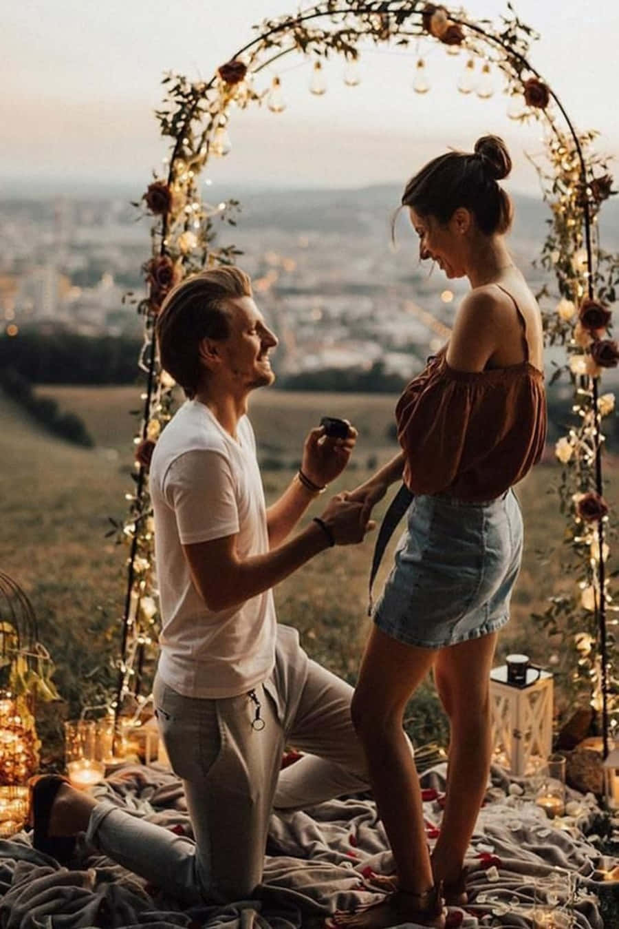 Proposal 桌布 壁紙 — 一名男子跪在花拱門下，向一位微笑的女子求婚，地點是俯瞰城市的小山頂，正值黃金時刻。