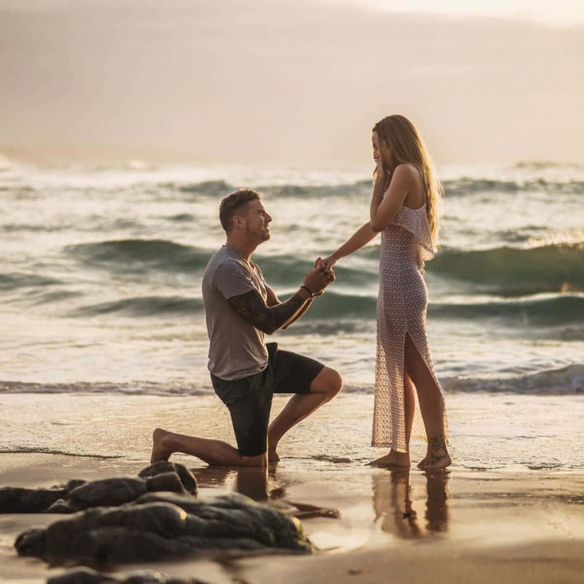 Proposal 桌布 壁紙 — 一名男子跪在陽光明媚的海灘上，向一位身穿印花裙的女子求婚，身後海浪翻滾。