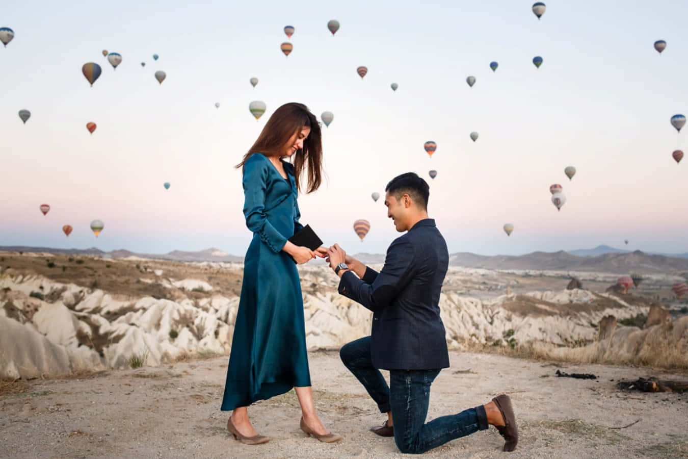 Proposal 桌布 壁紙 — 一名男子跪地向身穿青綠色連衣裙的女子求婚，黎明時分熱氣球漂浮在超現實的岩石景觀上空。