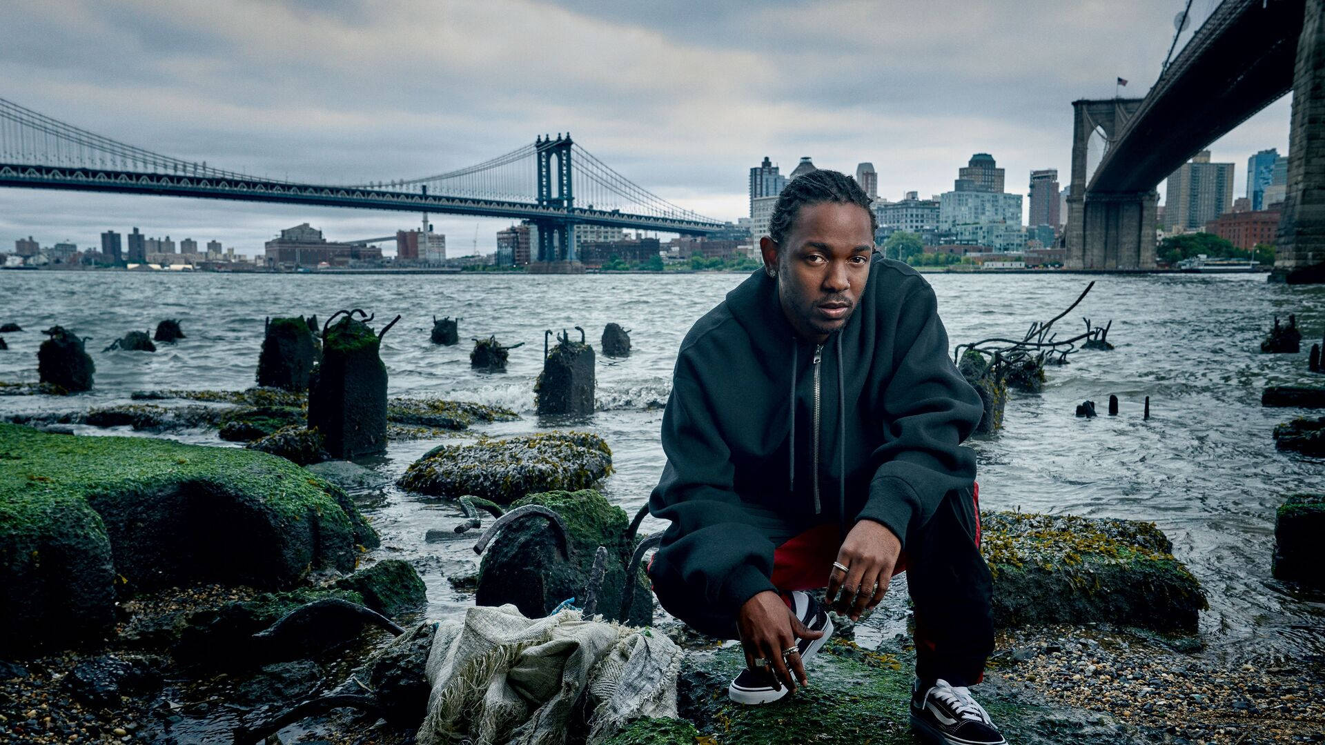Kendrick 桌布 壁紙 — 一名男子蹲在長滿苔蘚的岩石上，靠近水邊，背景是布魯克林大橋和城市天際線，天空多雲。