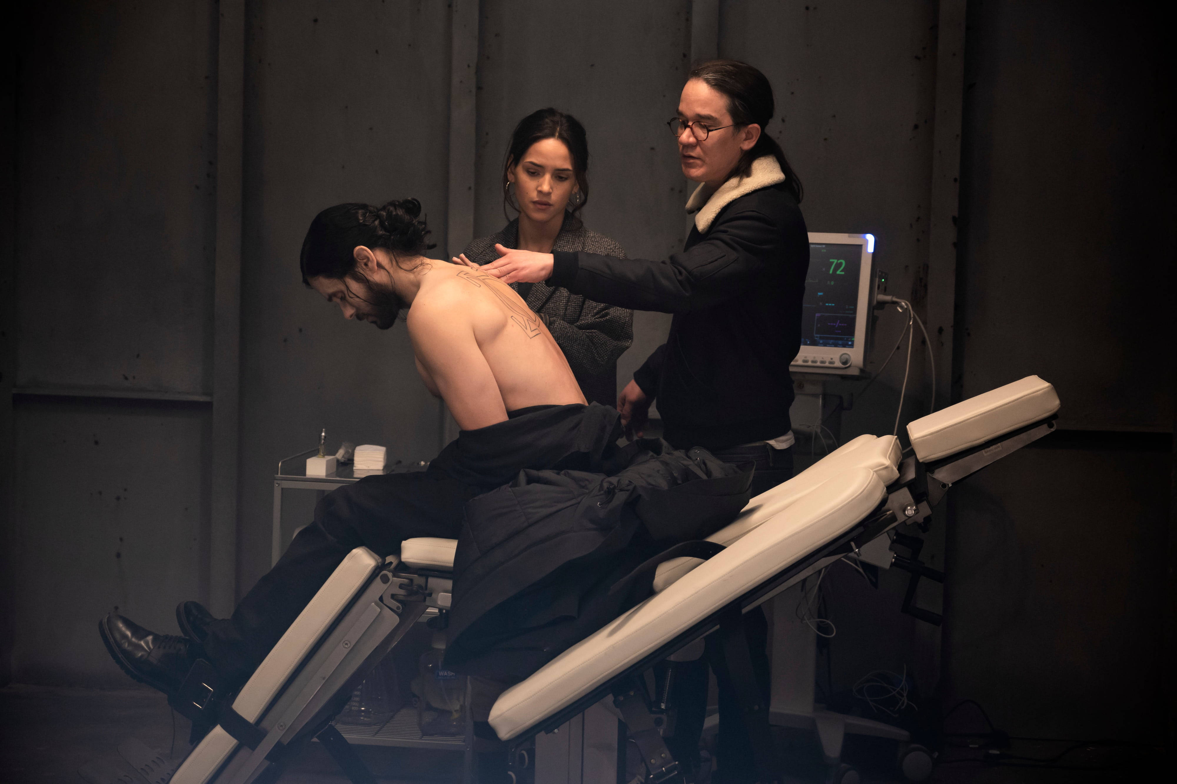 Morbius 桌布 壁紙 — 一名男子躺在醫療床上，兩名專業人員在昏暗的臨床房間檢查他的背部。
