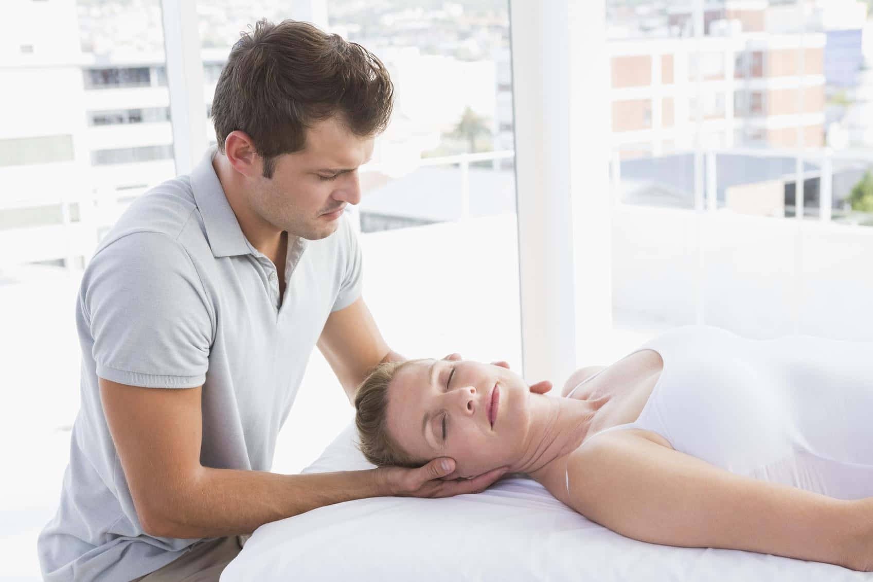 Massage 桌布 壁紙 — 一名男子輕柔地按摩一名女子的頸部，她則在白色靠墊上放鬆，旁邊是明亮的窗戶，窗外可見城市建築。