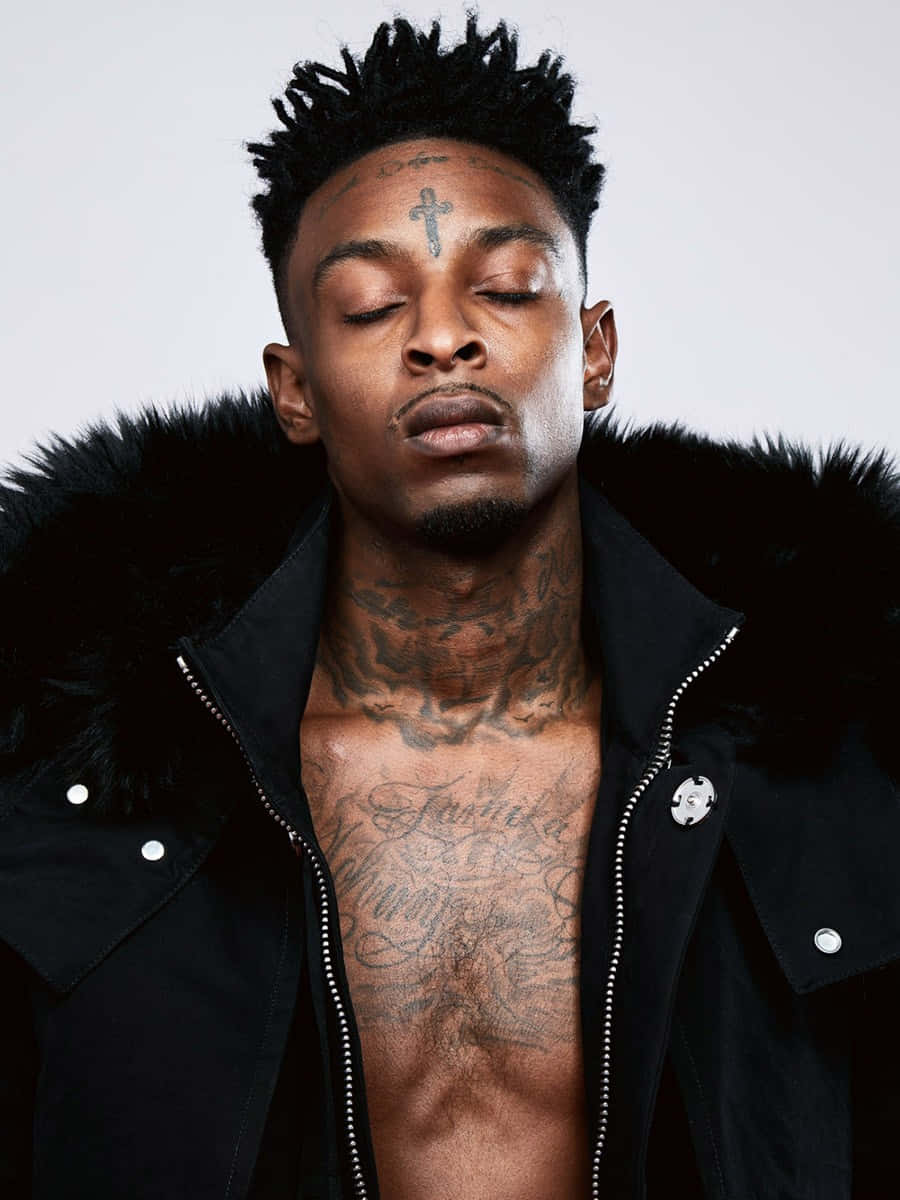 21 Savage 桌布 壁紙 — 一名男子閉上眼睛，胸前和額頭上有刺青，穿著黑色毛領外套，背景簡潔。