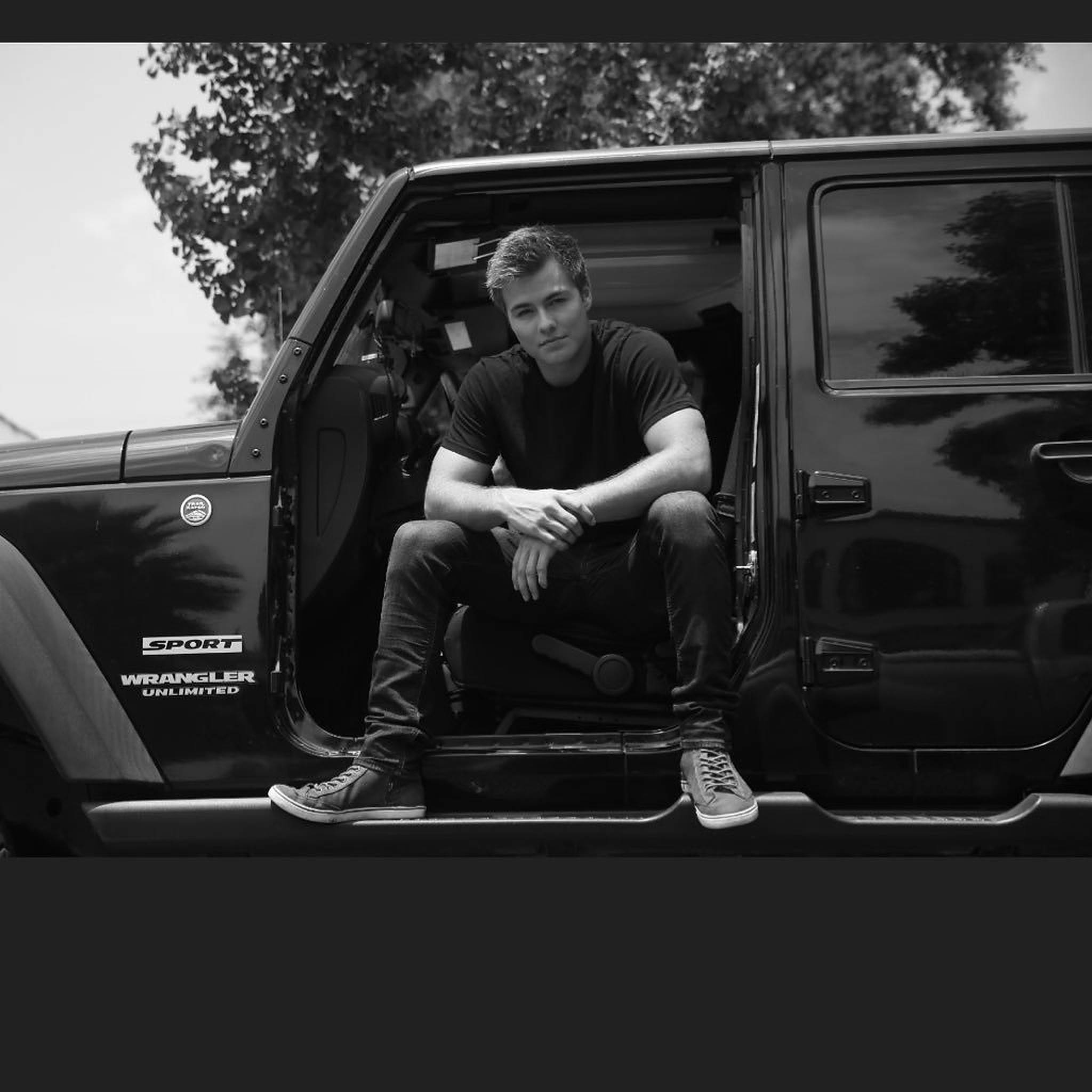 Peyton 桌布 壁紙 — 一名男子隨意地坐在黑色Jeep Wrangler Unlimited的敞開車門中，背景是樹木和天空。