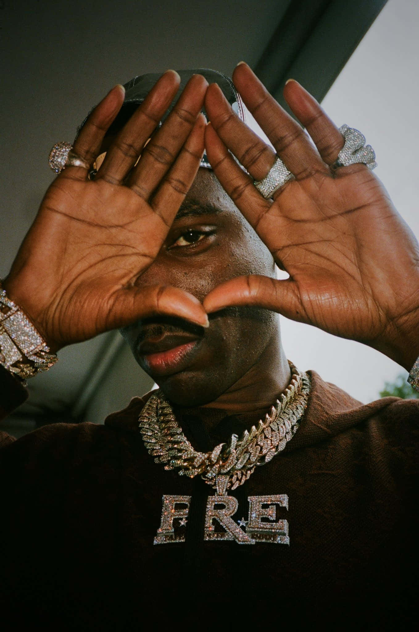 Young Dolph 桌布 壁紙 — 一名男子雙手托腮，佩戴鑲鑽首飾，身穿印有「FRE」字樣的連帽上衣，專注地凝視鏡頭。