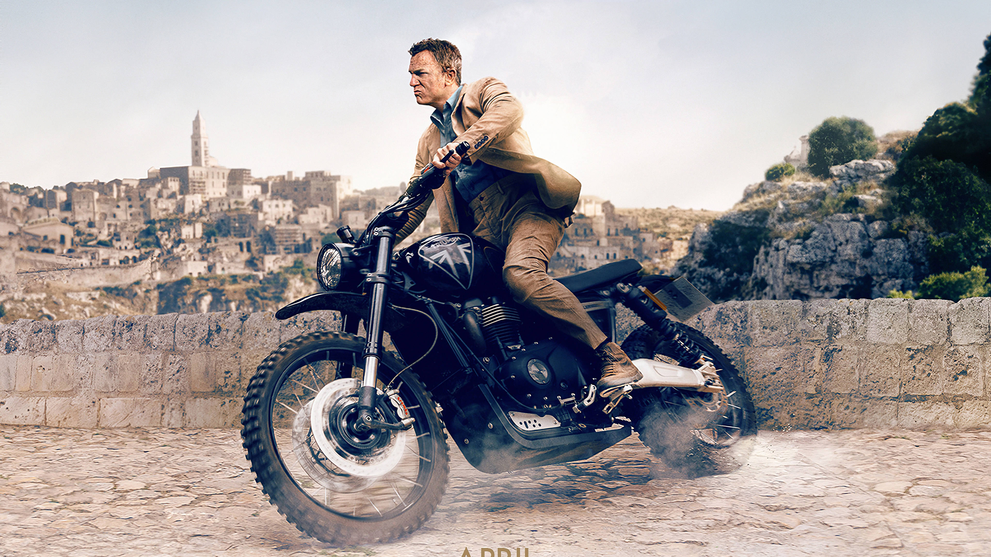 Daniel Craig 桌布 壁紙 — 一名男子騎著黑色摩托車在鵝卵石路上，背景是歷史悠久的石頭城鎮。