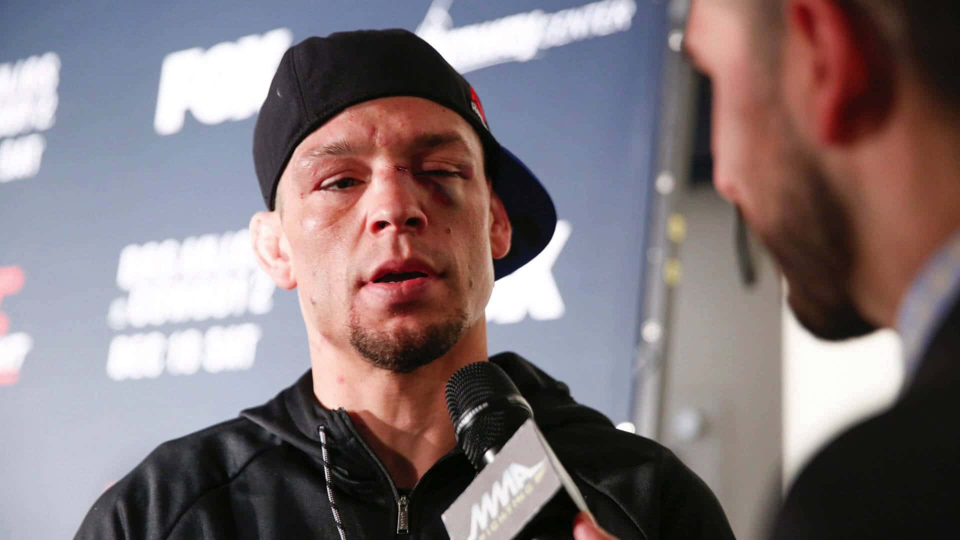 UFC 桌布 壁紙 — 一名男性MMA拳手戴著黑色帽子，眼睛瘀傷，在記者會上對著MMA品牌麥克風發言。