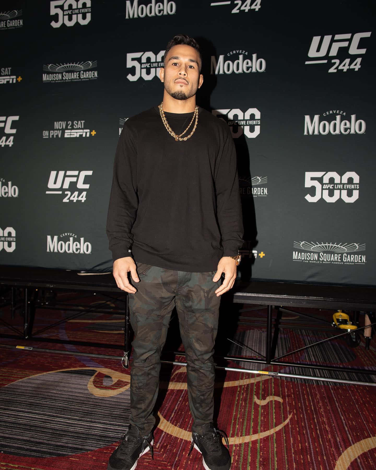 MMA 桌布 壁紙 — 一名男性拳手在麥迪遜廣場花園自信地站在UFC 244品牌背景前，身穿黑色運動衫和迷彩褲。