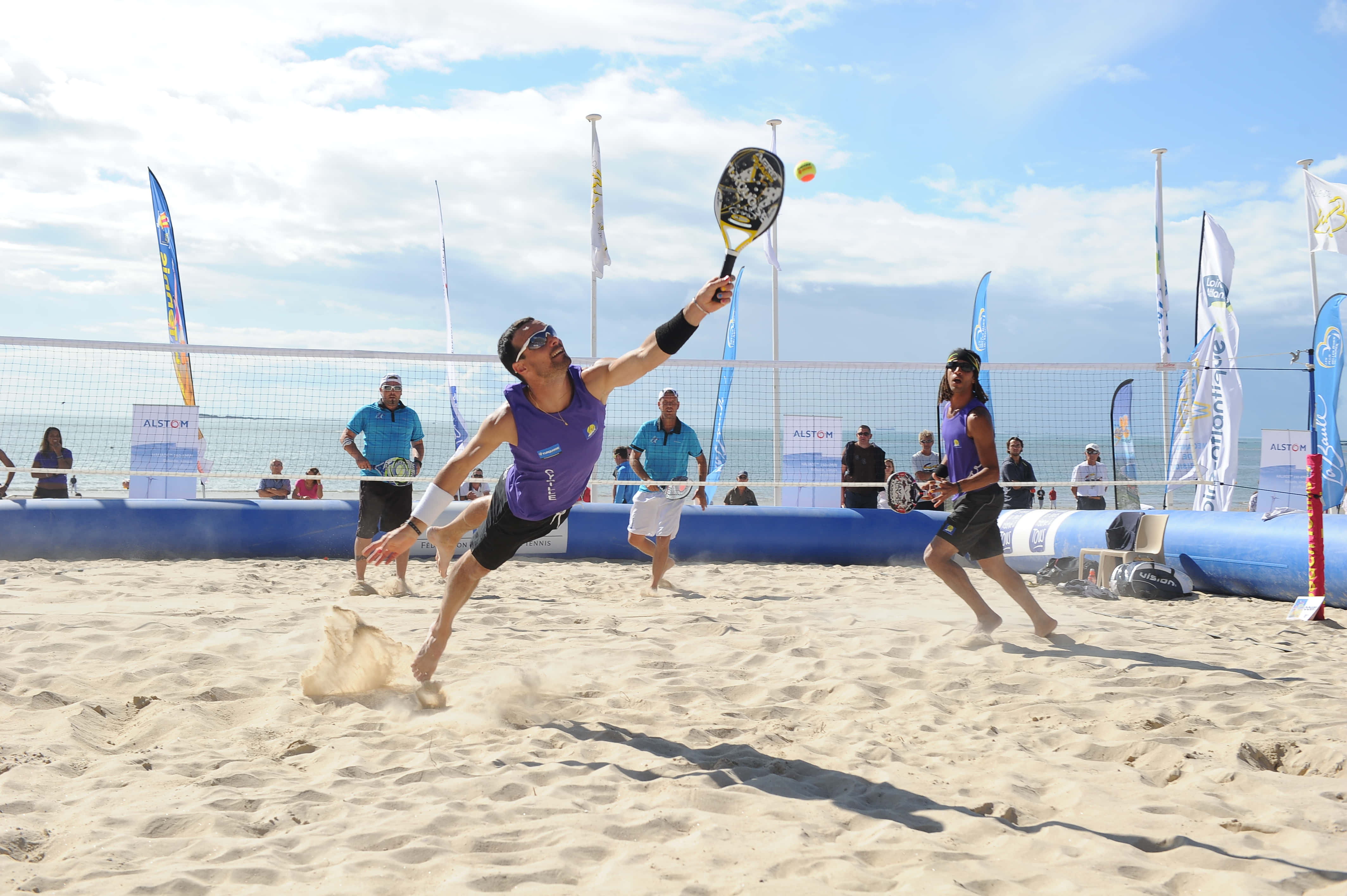 Beach Tennis 桌布 壁紙 — 一名男性沙灘網球選手動態跳起扣殺球，而他的隊友在陽光照耀的沙灘上觀看。
