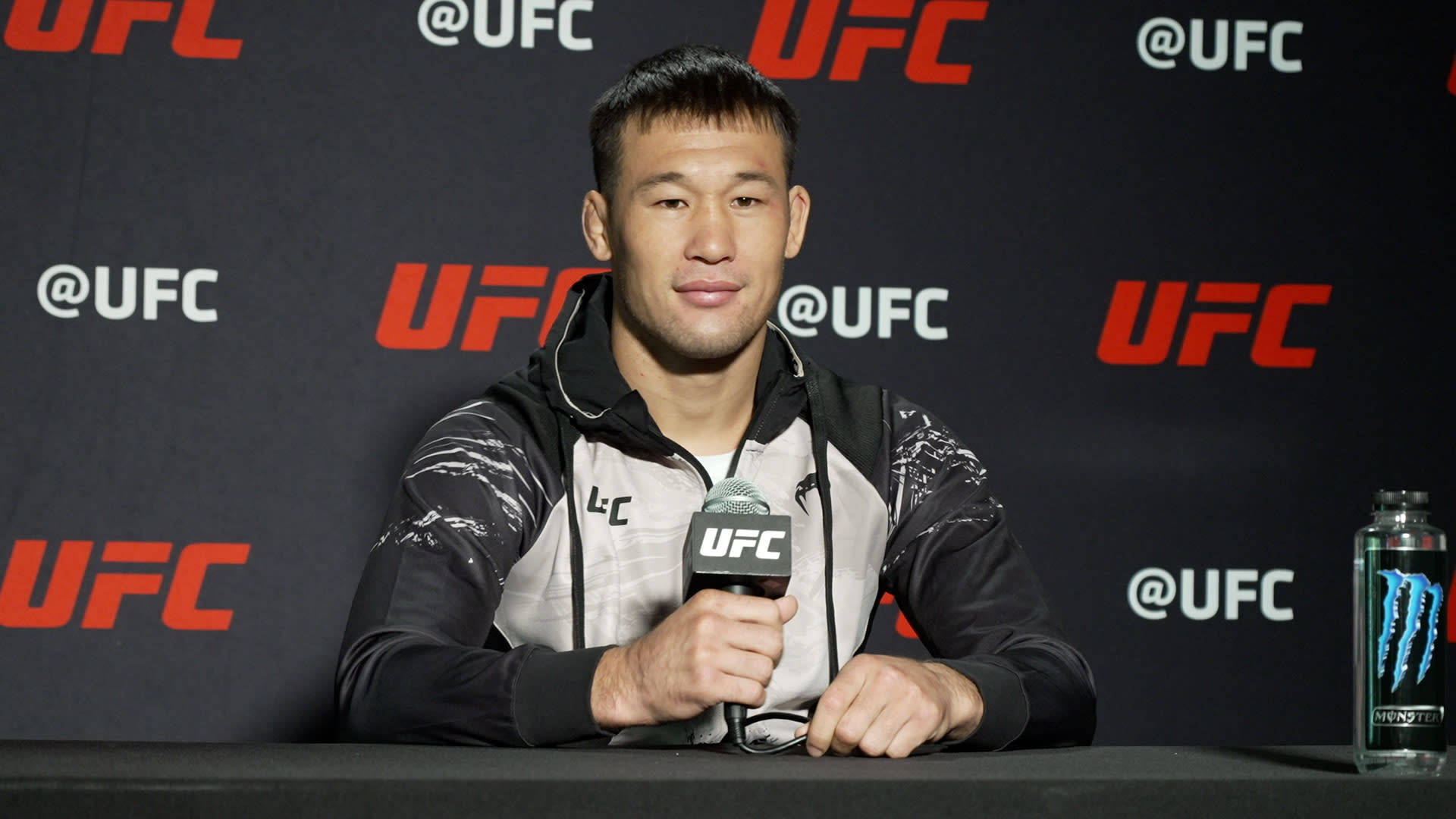 UFC 桌布 壁紙 — 一名男性混合武術選手坐在新聞發布會桌前，手持UFC麥克風，旁邊放著一瓶Monster Energy。
