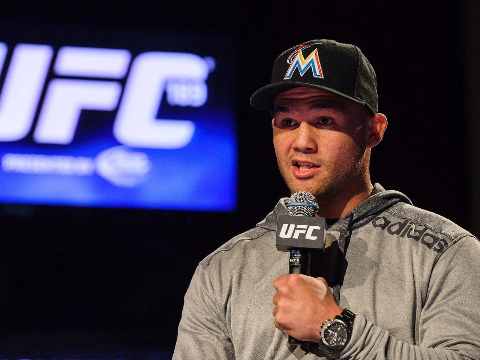 MMA 桌布 壁紙 — 一名男性綜合格鬥選手戴著邁阿密馬林魚帽和灰色連帽衫，對著UFC麥克風說話。