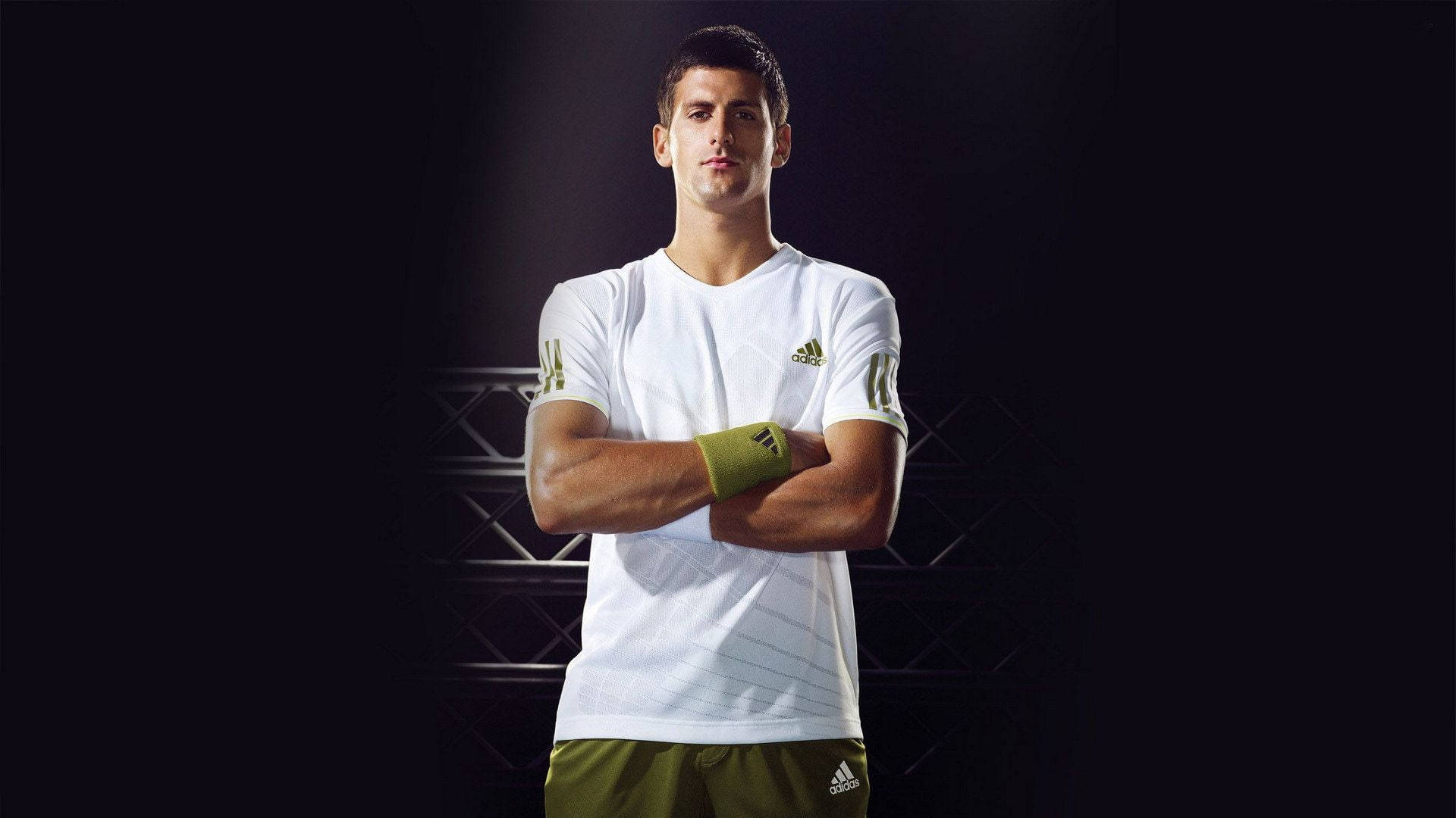 Novak 桌布 壁紙 — 一名男性網球運動員自信地交叉雙臂站在黑暗的攝影棚中，被聚光燈照亮，身穿白色和綠色的運動裝備。