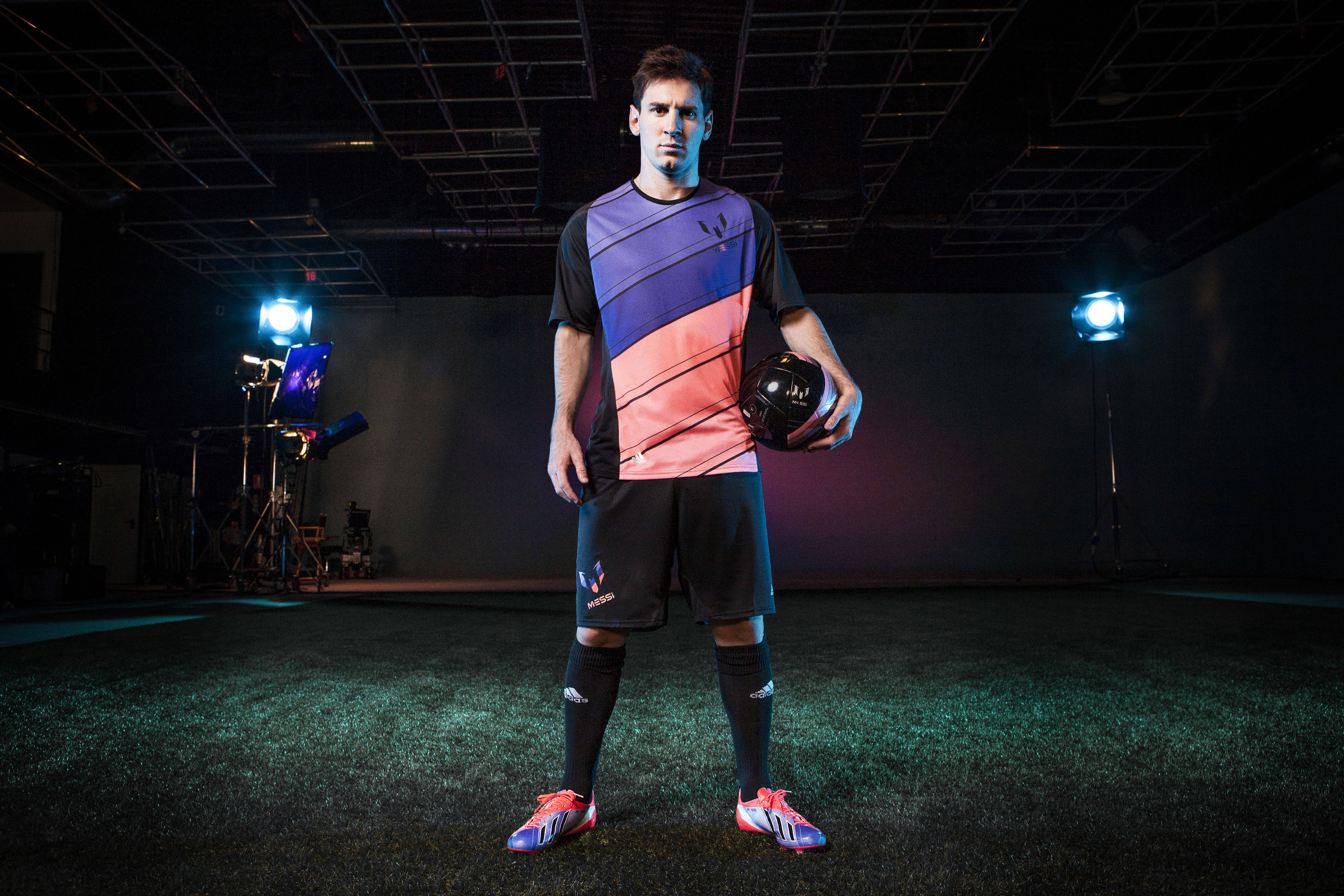 Messi 4K Ultra HD 桌布 壁紙 — 一名男性足球運動員穿著鮮豔裝備，在工作室燈光下自信地站在人造草皮上，手持足球。