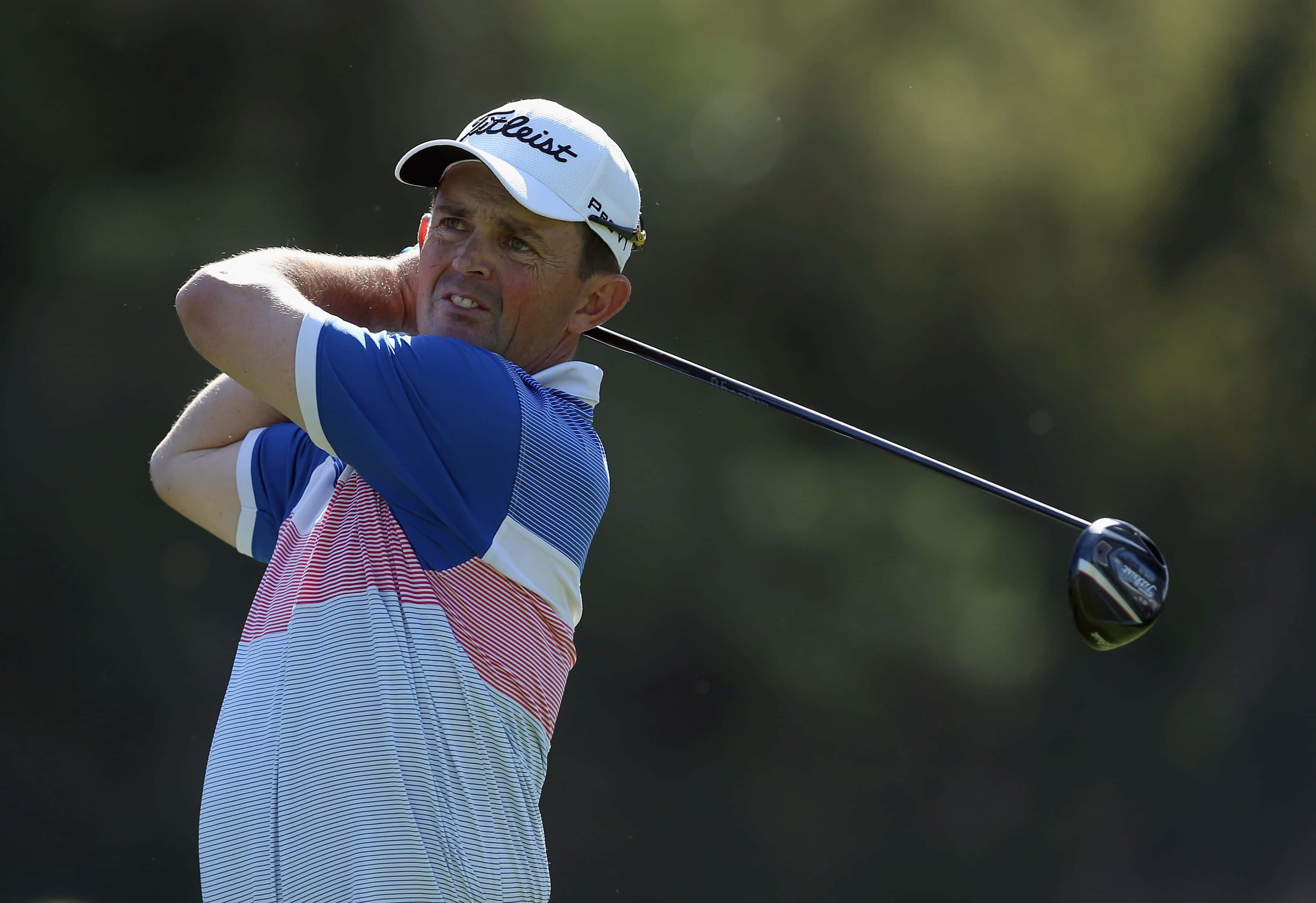 運動 桌布 壁紙 — 一名男性高爾夫球手在陽光明媚的球場上揮桿，背景有模糊的綠色樹葉。