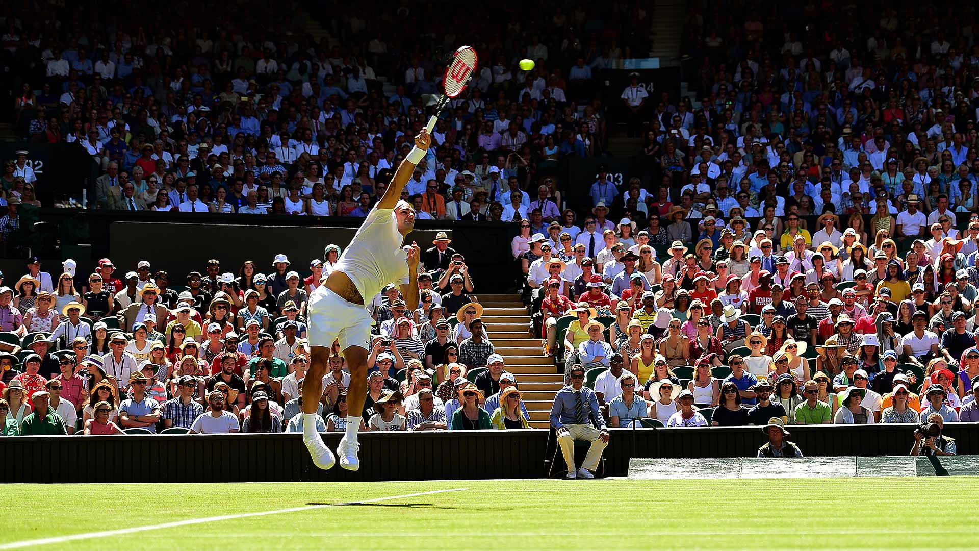 Wimbledon 桌布 壁紙 — 一名男網球選手在草地場上的比賽中，在明亮的日光下於座無虛席的體育館內高高躍起擊出正手拍。