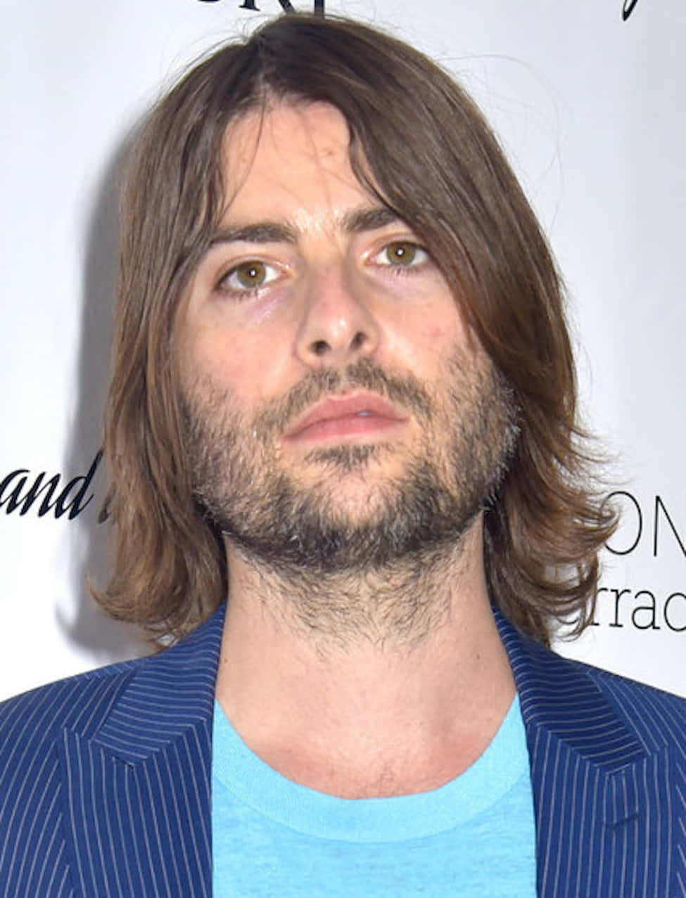 Jason Schwartzman 桌布 壁紙 — 一名留著及肩棕色頭髮和鬍鬚的男子，身穿藍色細條紋西裝外套，內搭淺藍色T恤，背景為白色，帶有模糊文字。