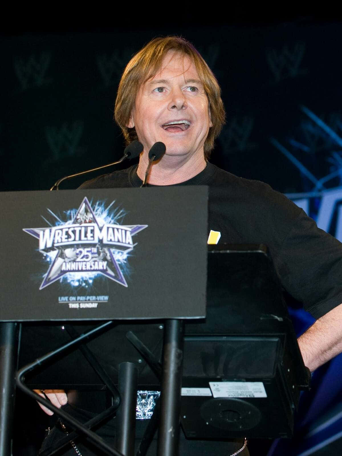 摔角 桌布 壁紙 — 一名留著及肩長髮的男子在舞台燈光下，於WrestleMania 25週年紀念講台上熱情地發言。
