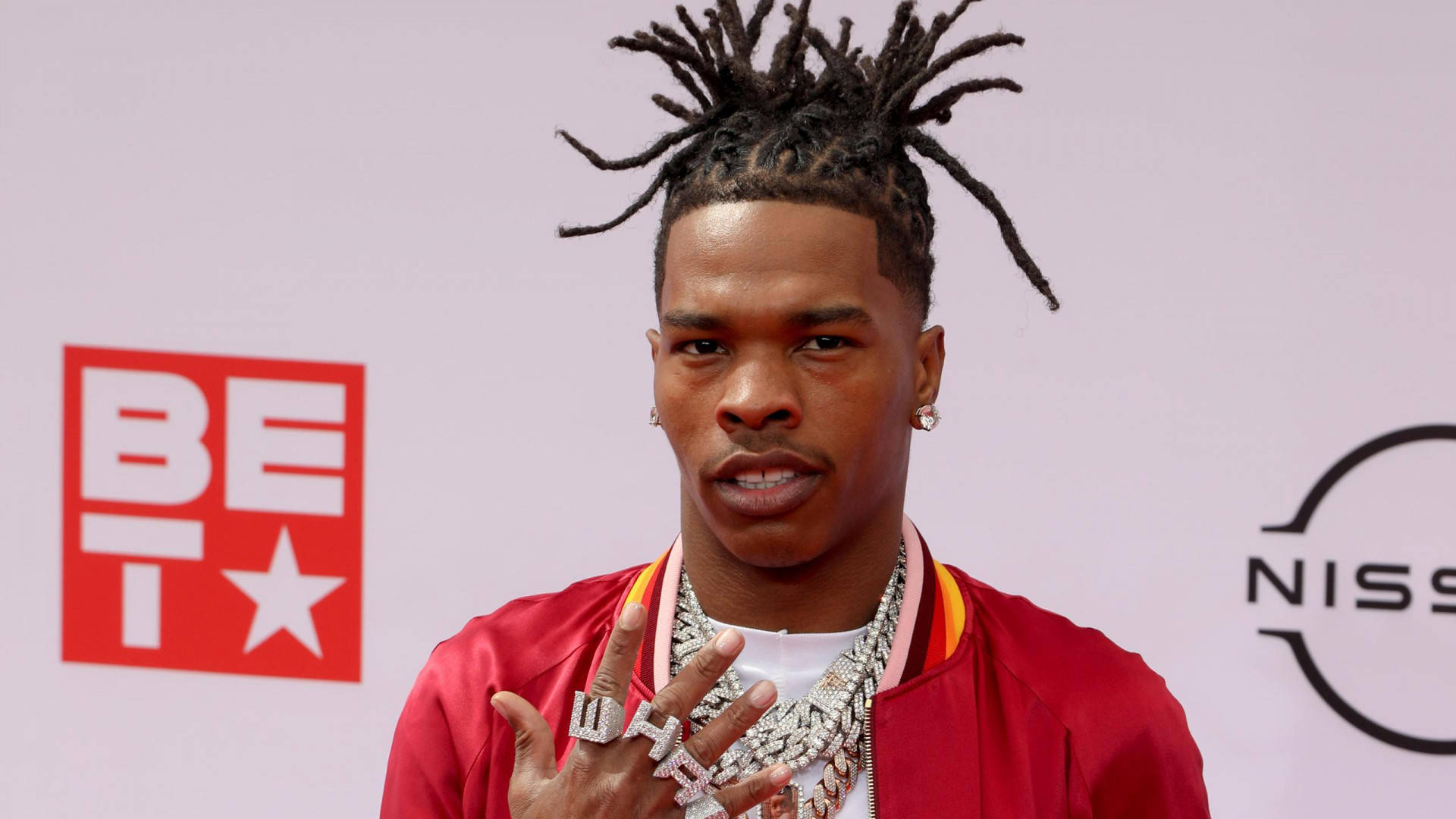 Lil Baby 桌布 壁紙 — 一名留著尖刺辮子頭的男子，身穿紅色外套和閃亮首飾，在身後有BET和日產標誌的紅地毯上擺姿勢。