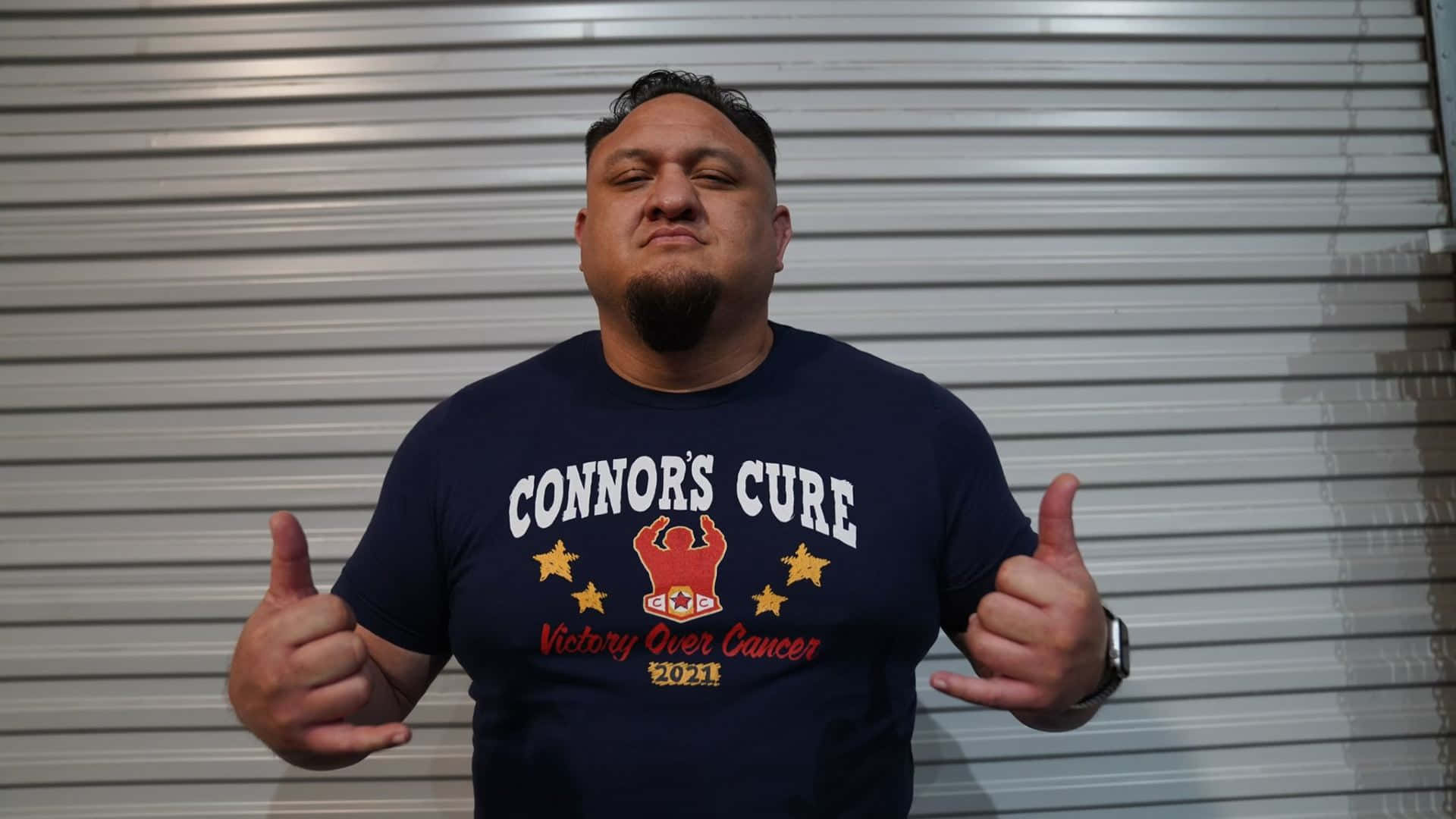 WWE 桌布 壁紙 — 一名留著山羊鬍的男士，身穿海軍藍T恤推廣Connor's Cure Victory Over Cancer 2021，在銀色捲門前豎起雙拇指。