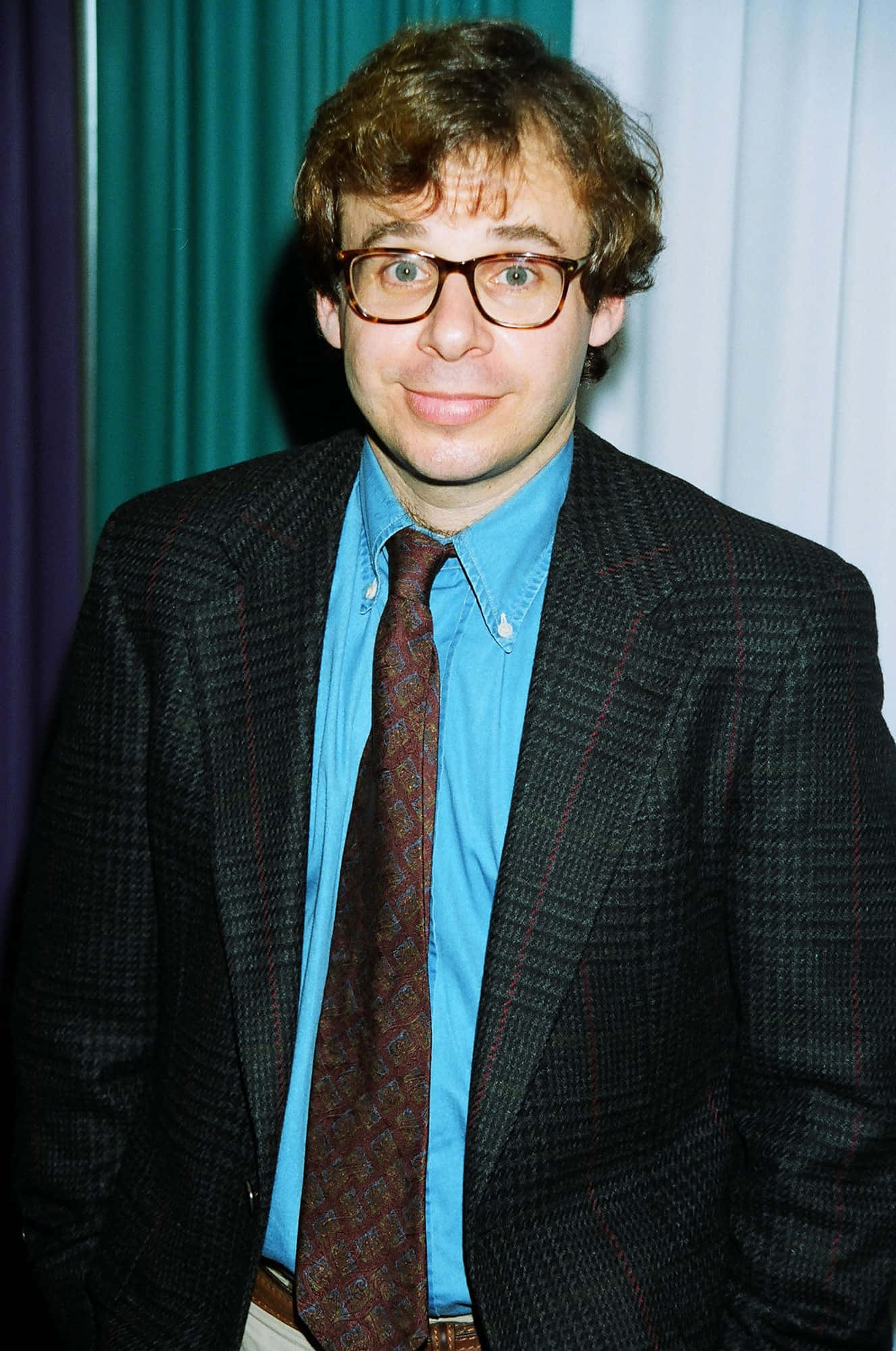 rick moranis 桌布 壁紙 — 一名留著捲髮、戴眼鏡的男子，身穿格紋西裝和圖案領帶，背景是藍綠色和白色的窗簾。